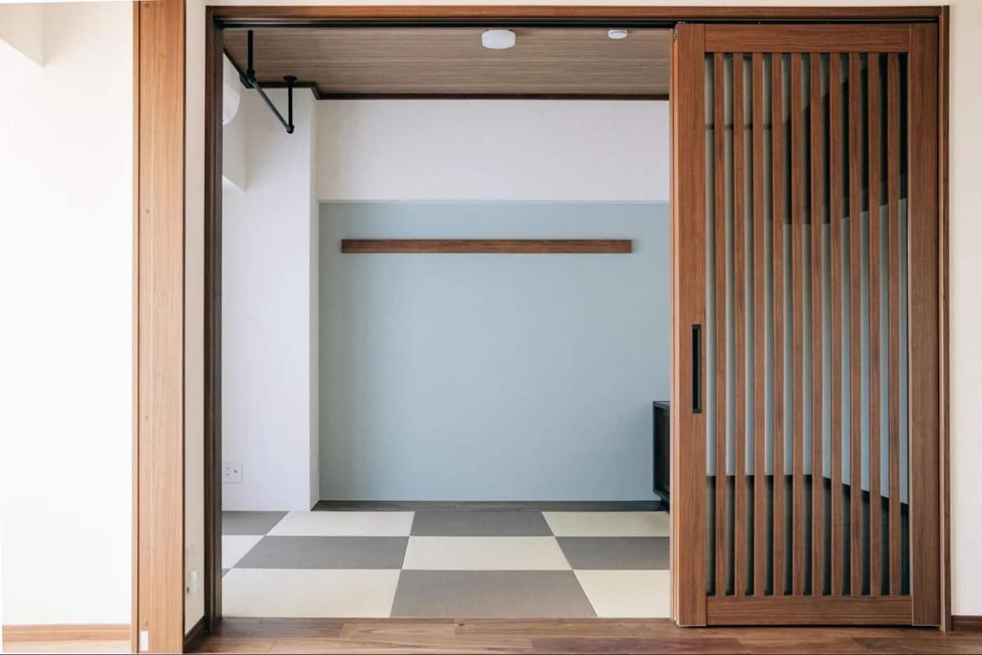 縦格子の木製引き戸と市松模様の床が特徴の和モダン空間。水色のアクセント壁に水平の木製梁、木天井に円形照明と黒のアイアンバーを備える。