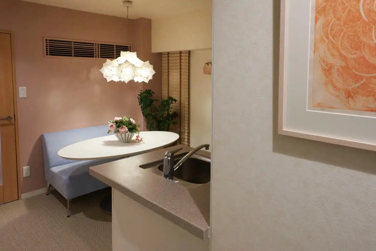 ダイニングキッチンの写真。手前にキッチンシンク、奥に青いベンチと白いテーブルが置かれている。壁はピンク色で、白いペンダントライトが吊り下げられている。