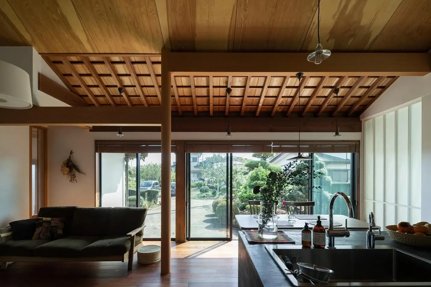 格子状の現し天井と大きな掃き出し窓があるLDK。手前のモルタル調キッチン越しに庭の緑を望む、古民家の趣とモダンなインテリアが調和した開放的な内観。