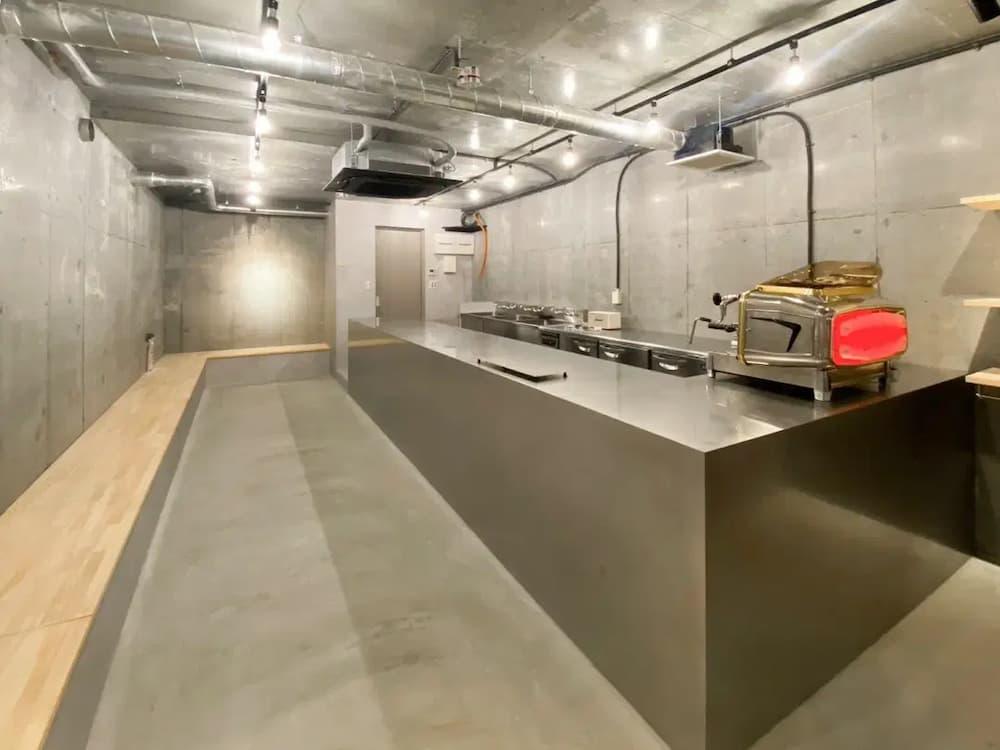 全面コンクリート打ちっ放しの壁と天井、グレーのモルタル床で構成された無機質な空間。中央には重厚なオールステンレス製のロングカウンターとシンクを配置し、赤いエスプレッソマシンが視線を引く。天井には銀色の露出配管や裸電球が走り、左手の木製ベンチが冷たさを和らげる、倉庫をリノベーションしたようなインダストリアルテイストのキッチンの内観