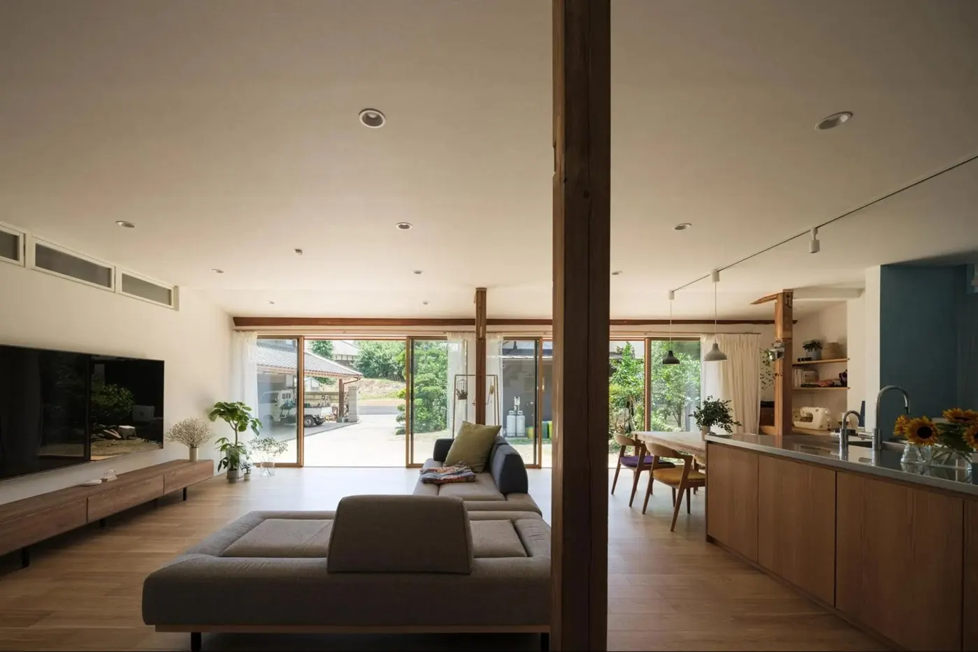 築50年の古民家をリノベーションした、庭と繋がる開放的なLDK。既存の柱や欄間を残しつつ、続き間の和室を現代的な大空間に改修。木製の対面キッチンとグレーのソファを配置し、親戚が集まれる広さを確保した株式会社AS IT ISの事例。