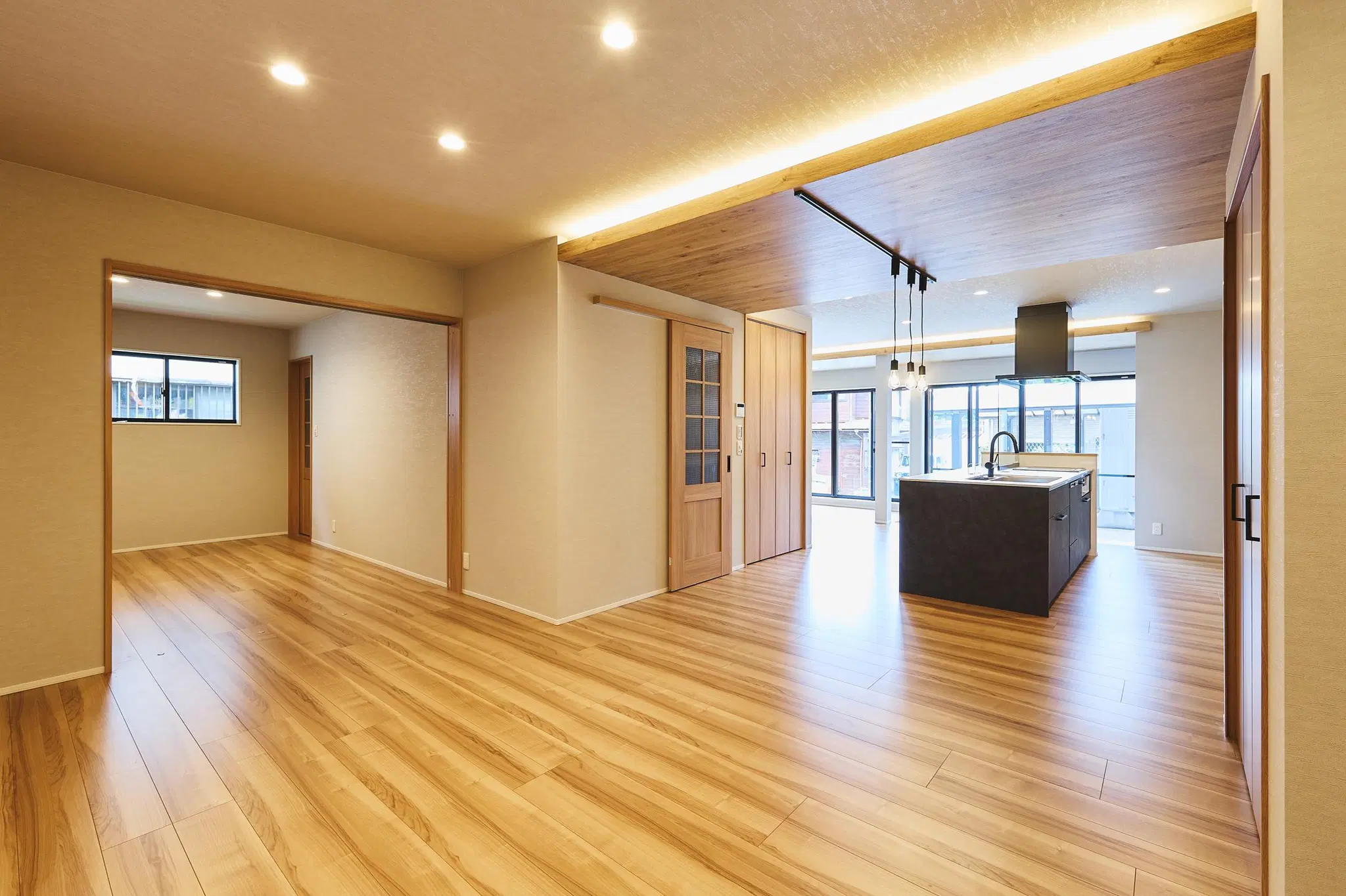 実家をフルリノベーションした戸建ての広々としたLDK。温かみのある木目調フローリングが広がる空間。右手には重厚感のあるブラックのアイランドキッチンが配置されており、その上部は間接照明を仕込んだ木目調の下がり天井になっている。黒と木目のコントラストが美しく調和したモダンな室内。奥には外の自然光を取り込む大きな窓が連続して設けられている。My Design(株式会社Izumida)のリノベーション事例。