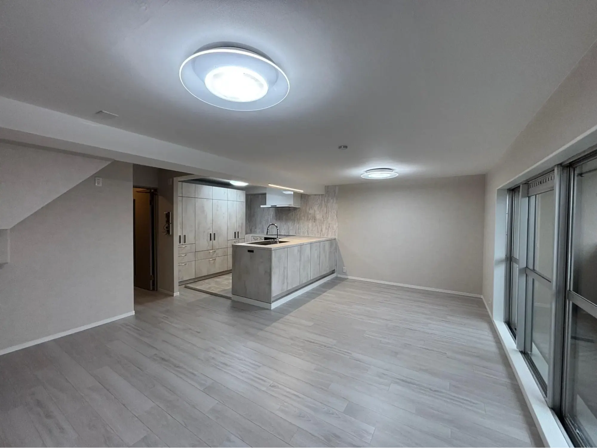 リノベーション後のLDK。ライトグレーのペニンシュラキッチンと大容量の背面収納を配置。床はアッシュ系の白木目フローリング。右手に大きな窓があり、白とグレーで統一された明るくモダンな空間。吾妻工務店のリノベーション事例。