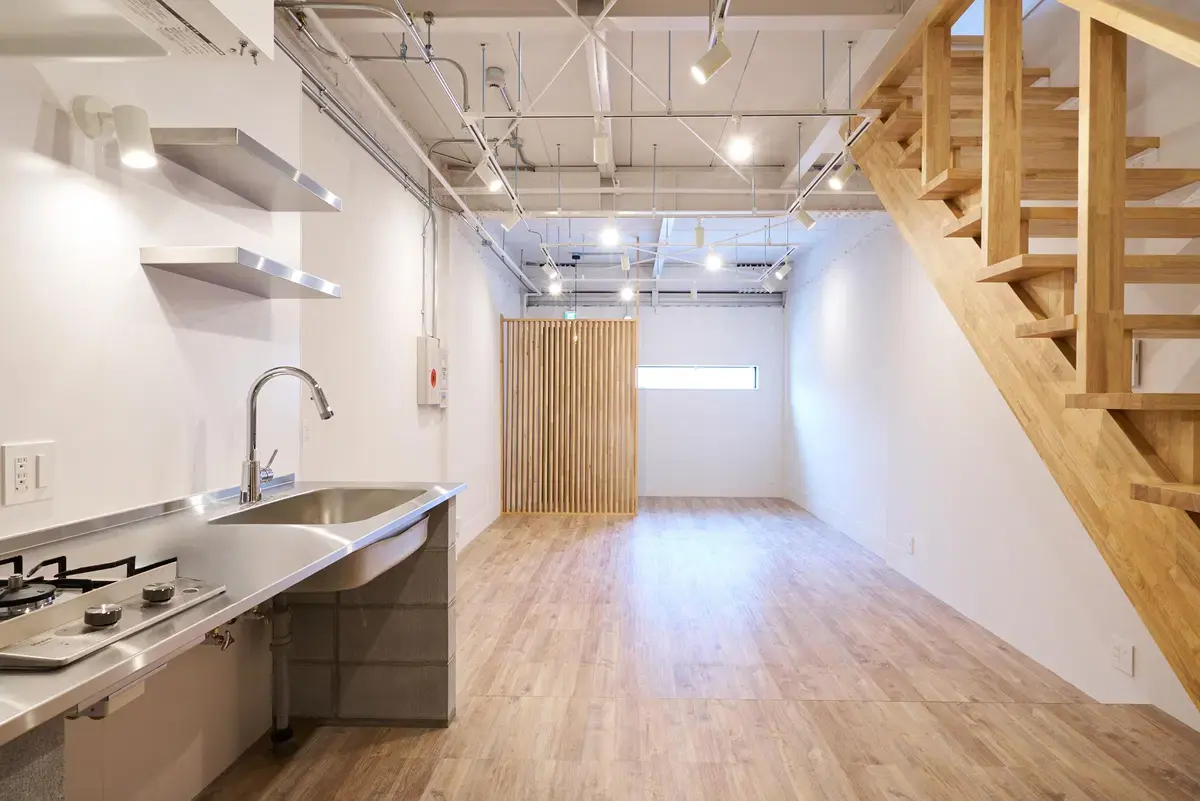 左側にステンレスフレームキッチンと壁面棚、右側に木製スケルトン階段を配置。白塗装の鉄骨剥き出し天井、配管、ダクトレール照明が特徴的な、明るく開放的な空間。ANRI STYLEのリノベーション事例。
