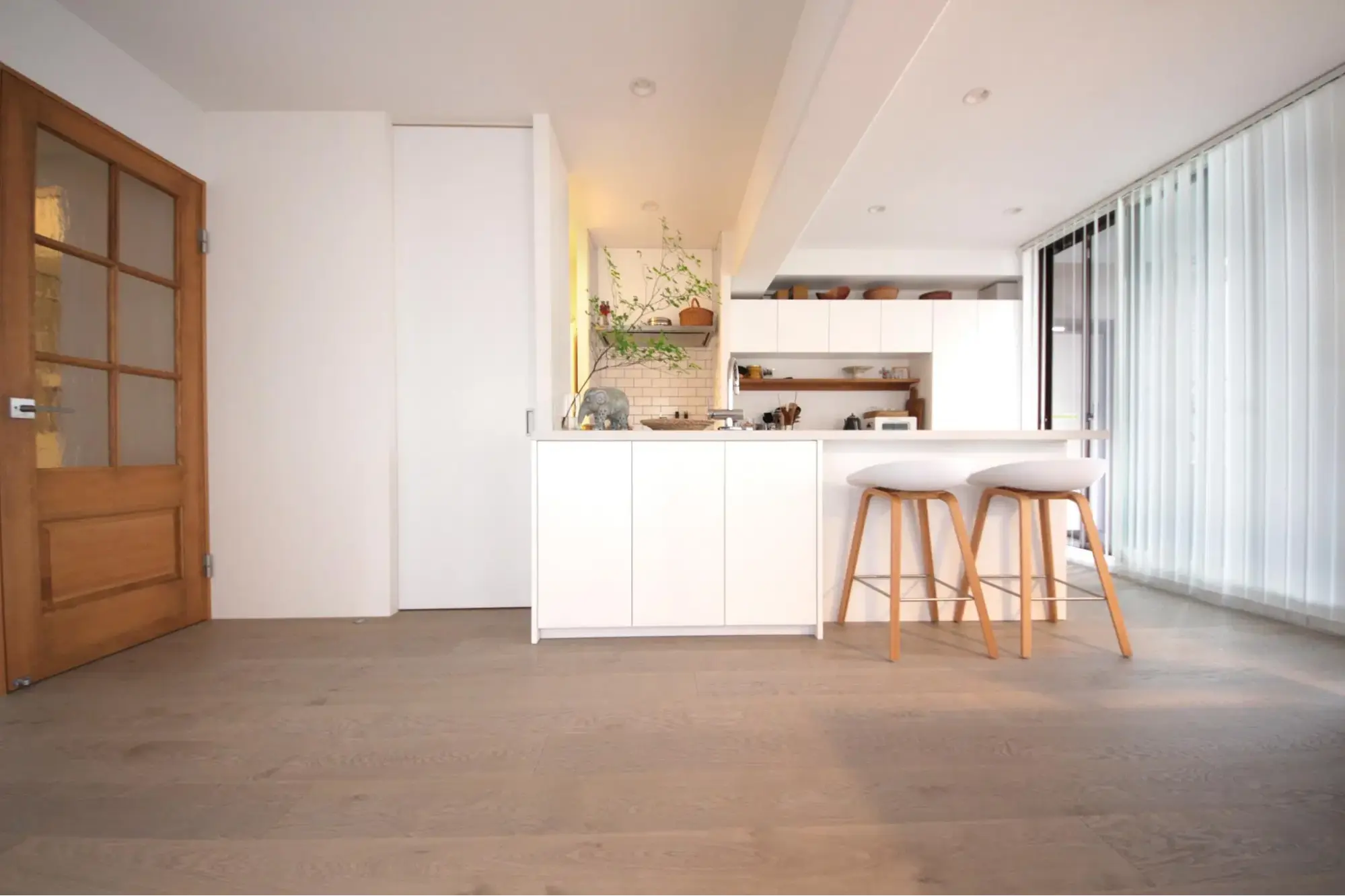 海外製の挽板フローリングと白い壁が明るく広々とした空間。中央にはシンプルでノイズレスなオーダーメイドの白いオープンキッチンを配置し、ハイスツールやガラス入りの木製ドアを合わせたナチュラルでカフェのようなマンションリノベーション。田中建設の施工事例。