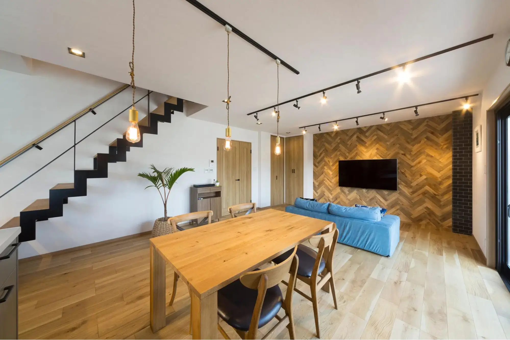 手前に木製ダイニング、左手に黒いアイアンのスケルトン階段を配置。奥にはブルーのソファと、テレビを掛けたヘリンボーン柄の壁面がある西海岸風のLDK。an cube（アン キューブ）のリノベーション事例。