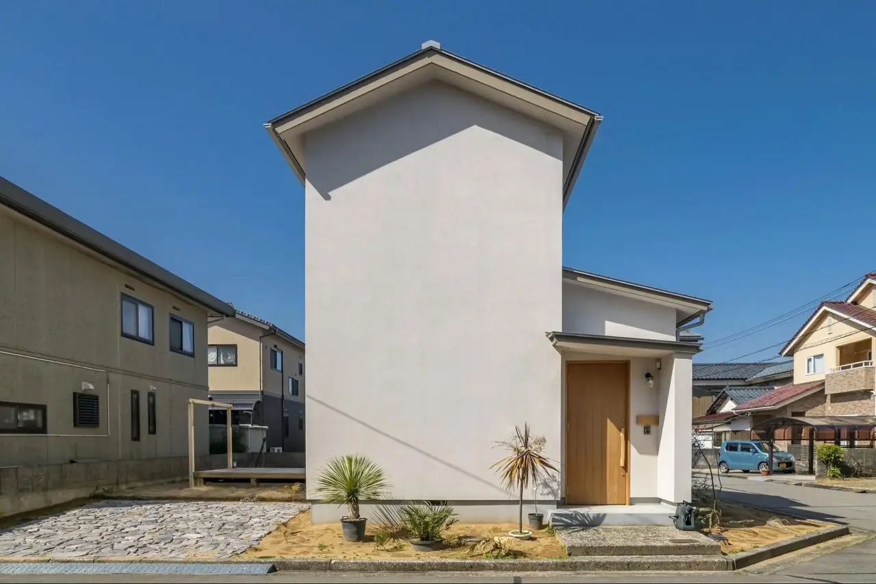 ベージュ系の塗り壁が特徴的な戸建て住宅の外観。シンプルな切妻屋根で、木製の玄関ドアと小さな植栽が配置されている。