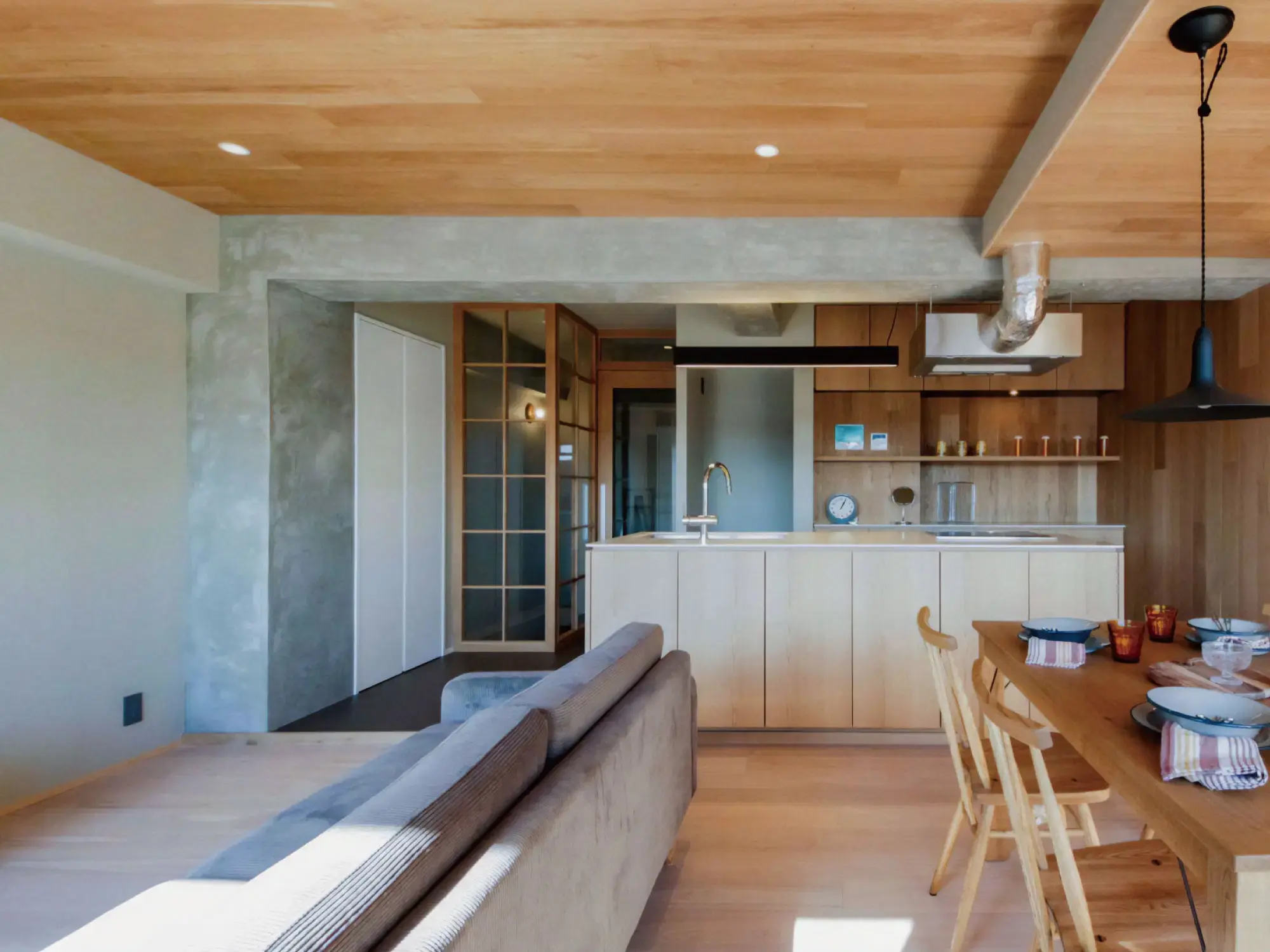 木の温もりを感じる板張りの天井と、無機質なモルタルの壁や柱のバランスが絶妙なナチュラルモダンなLDK。明るい日差しが差し込む空間に、木のアイランドキッチンやガラス入りの木製ドアを合わせ、居心地良く仕上げたマンションリノベーション。田中建設の施工事例。