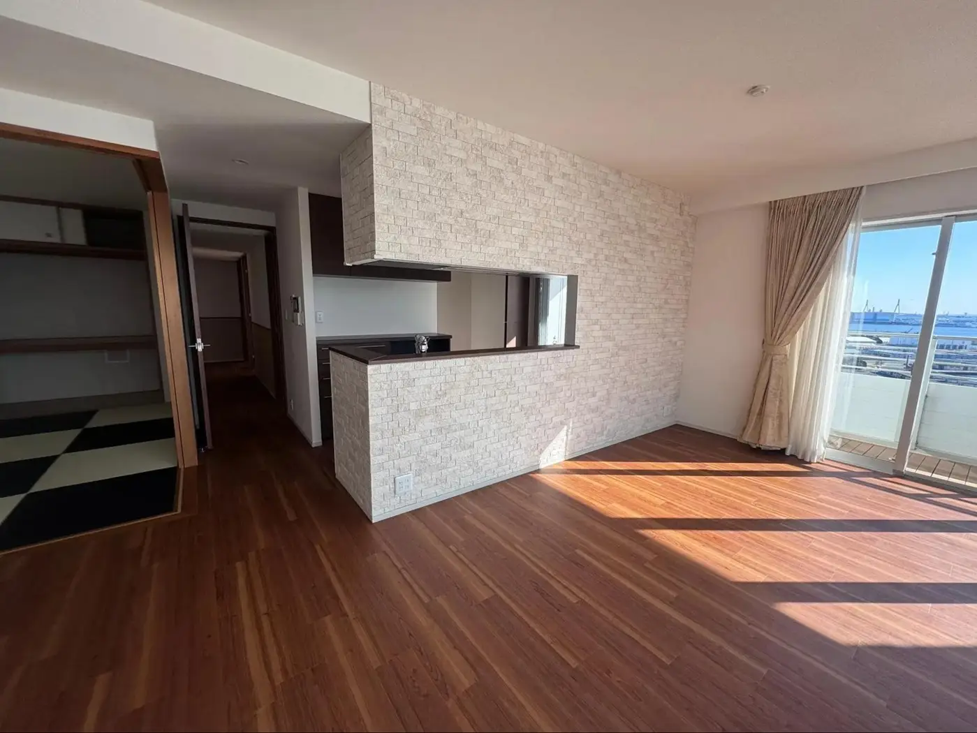 リフレネクスト横浜によるタワーマンションのリノベーション事例。石積み調のアクセントクロスで仕上げた対面キッチンの腰壁と垂れ壁が特徴的なLDK。リビングの大きな窓からは海や港の眺望が広がり、隣接する和室の様子も伺える。