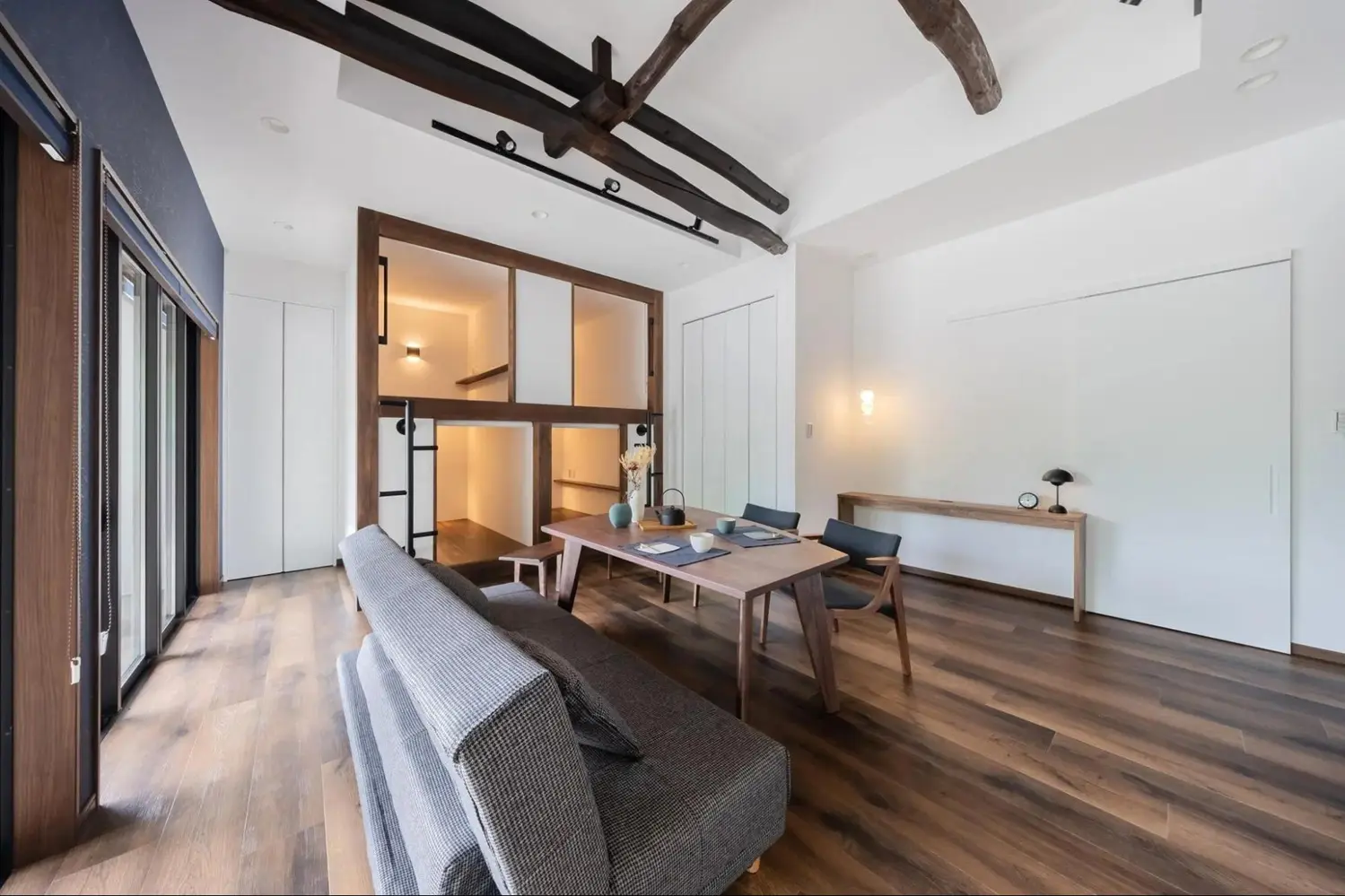 築100年の古民家を再生した、高い天井と太い梁が印象的なリビングダイニング。奥には梯子付きのカプセルホテル風ベッドスペースを設け、手前にはモダンな家具を配置してレトロモダンな空間に仕上げた民泊施設。ツクリカエの施工事例。