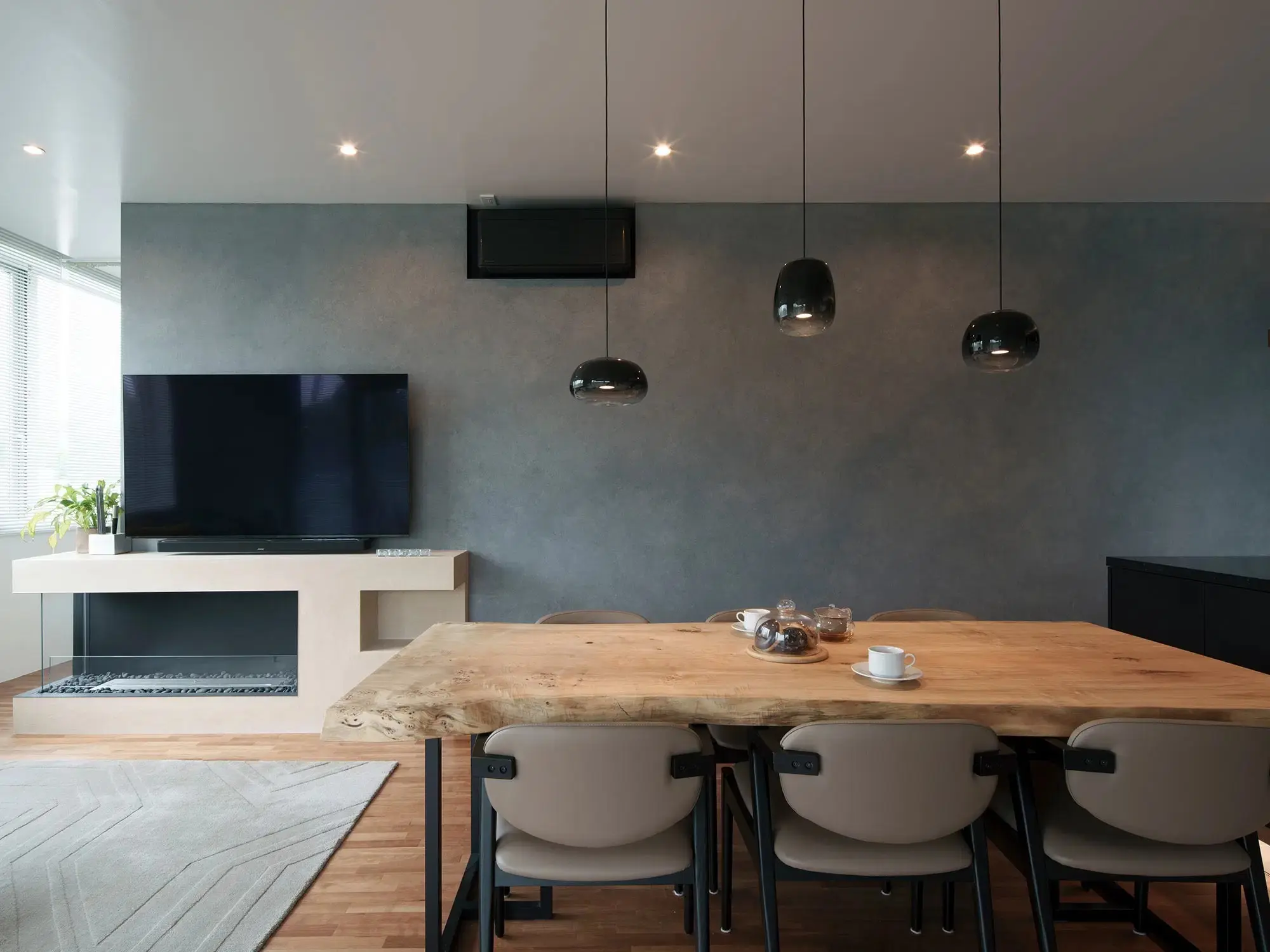 インテリックス空間設計のリノベーション事例。グレーの壁が印象的なホテルライクなダイニングには、一枚板の木製テーブルとモダンなチェアを配置。左手のテレビ下にはバイオエタノール暖炉が埋め込まれている。