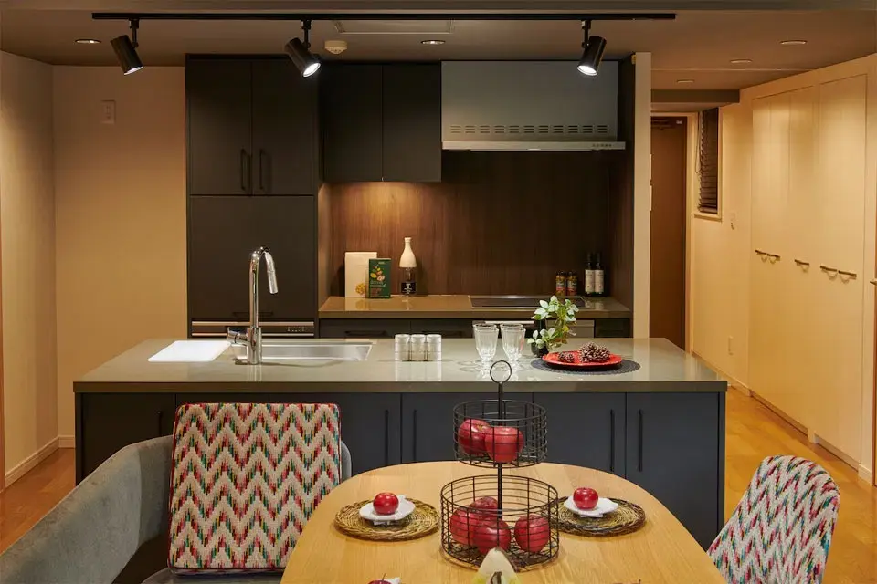 家の中心に配置された、ダークグレーのスタイリッシュなアイランド型オープンキッチン。背面の木目調パネルとスポットライトがホテルライクな空間を演出。手前には特徴的な柄のチェアを合わせ、家事をしながら家族の団らんを楽しめるマンションリノベーション。株式会社ディセノの施工事例。