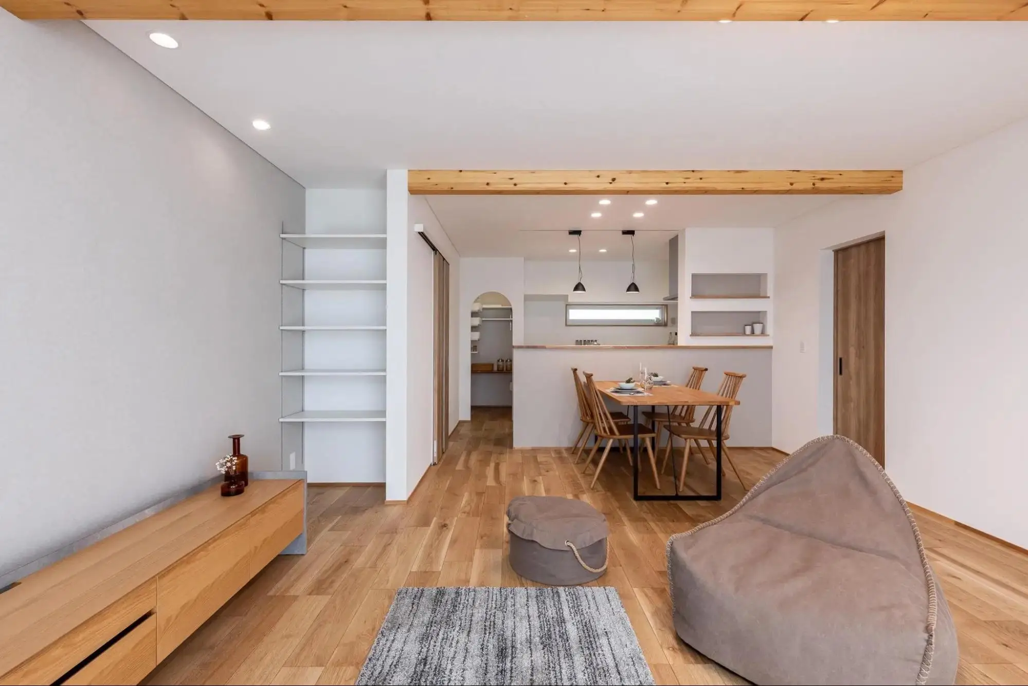 小林創建のリノベーション事例。天井の木の化粧梁がアクセントの、白と木目で統一されたナチュラルなLDK。奥にはキッチン横のアーチ壁や壁面収納が見え、手前はリビングスペースとなっている。