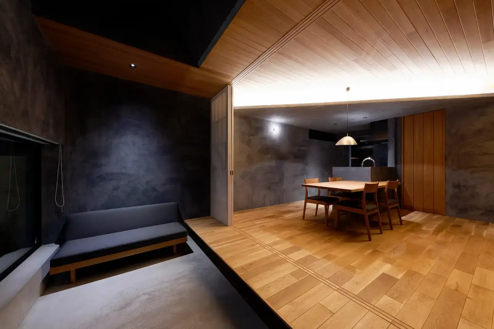 手前は一段下がったモルタルの土間空間でグレーのソファを配置。続く縁側風の木目床には木製ダイニングと奥にキッチンがある。板張りの天井と間接照明、ダークトーンの壁で構成された和モダンな空間。ひかり工務店のリノベーション事例。