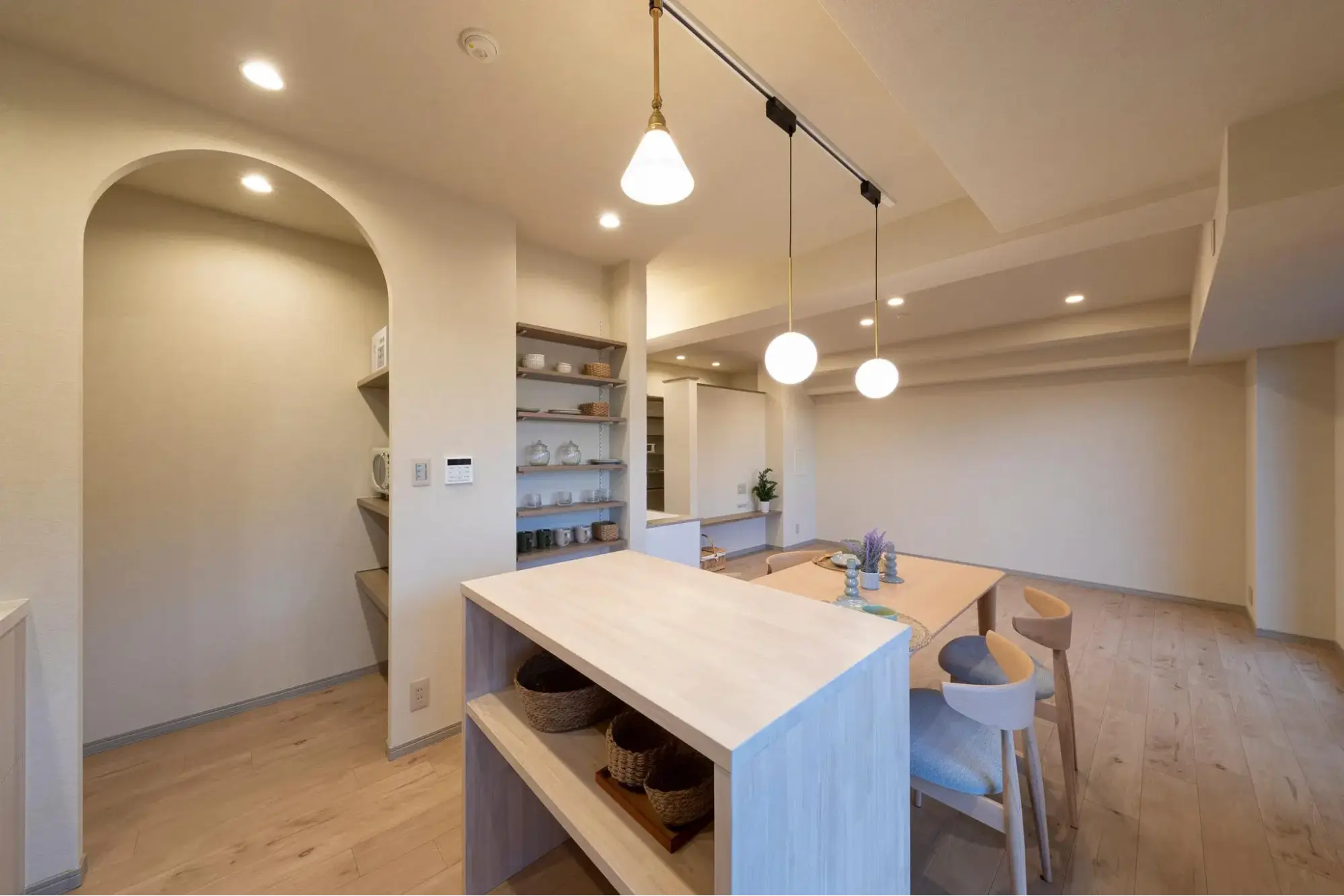 アーチ状の開口部を持つパントリーと、真鍮色のペンダントライトがアクセントのダイニングキッチン。手前には作業台兼収納カウンターを配置し、白と木目で統一した明るくホテルライクな空間。an cubeの施工事例。
