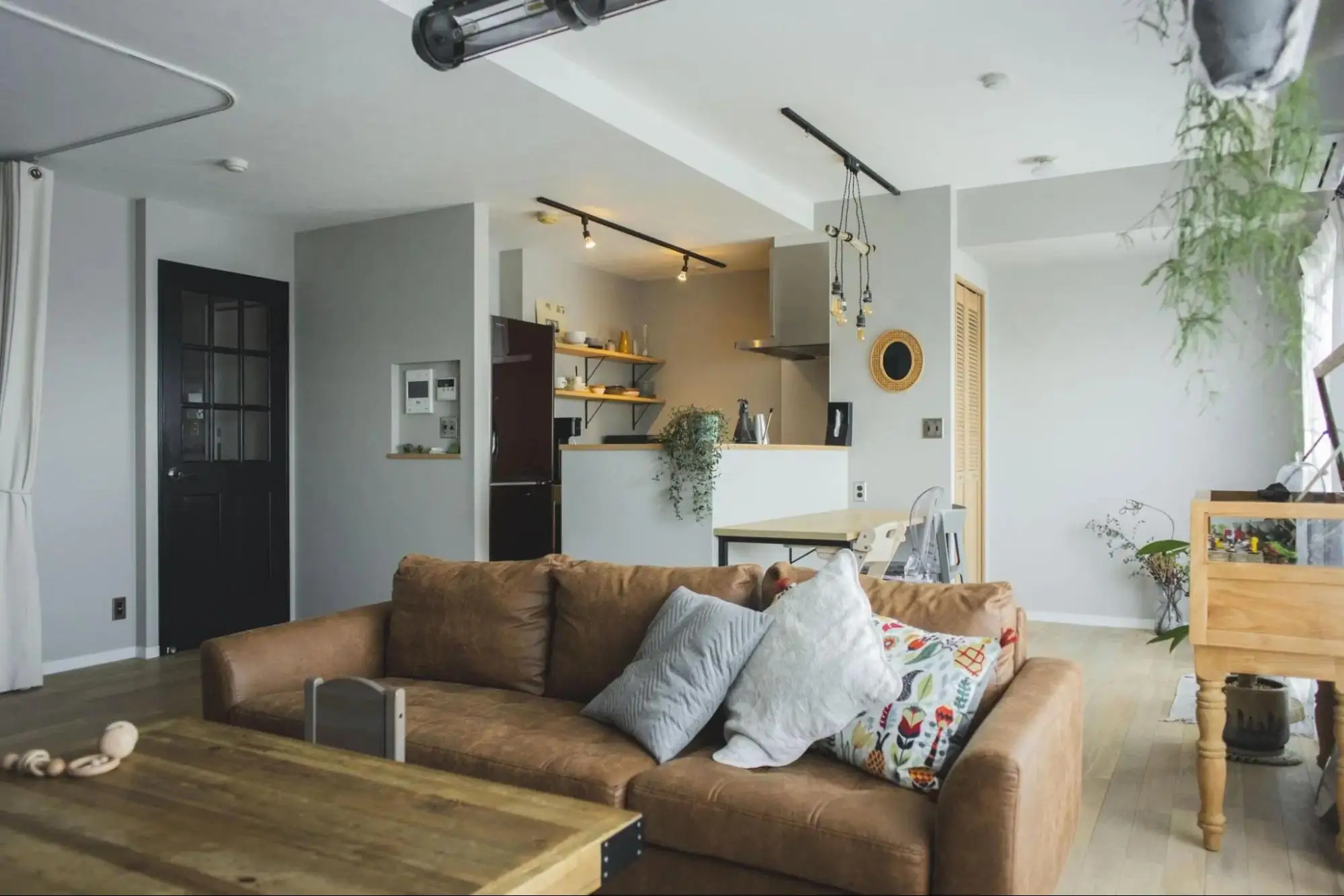 パパママハウスのリノベーション事例。淡いグレーの壁と黒いリビングドアが特徴的なLDK。手前には茶色のレザーソファ、奥には対面キッチンがあり、観葉植物や照明が飾られている。