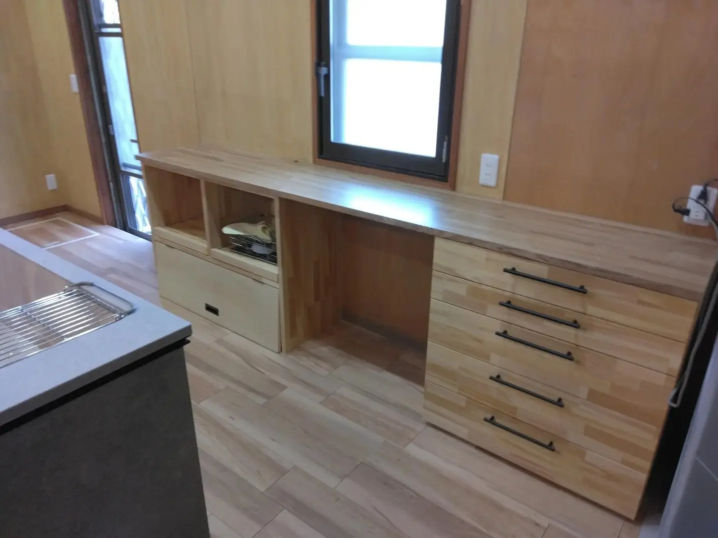 キッチンの背面に設置された、村山製材所による造作の木製カップボード。窓枠の下にぴったりと収まるよう設計され、黒い取っ手の引き出しとオープンスペースを備えている。無垢フローリングと調和する、機能的で温かみのあるオーダーメイド収納