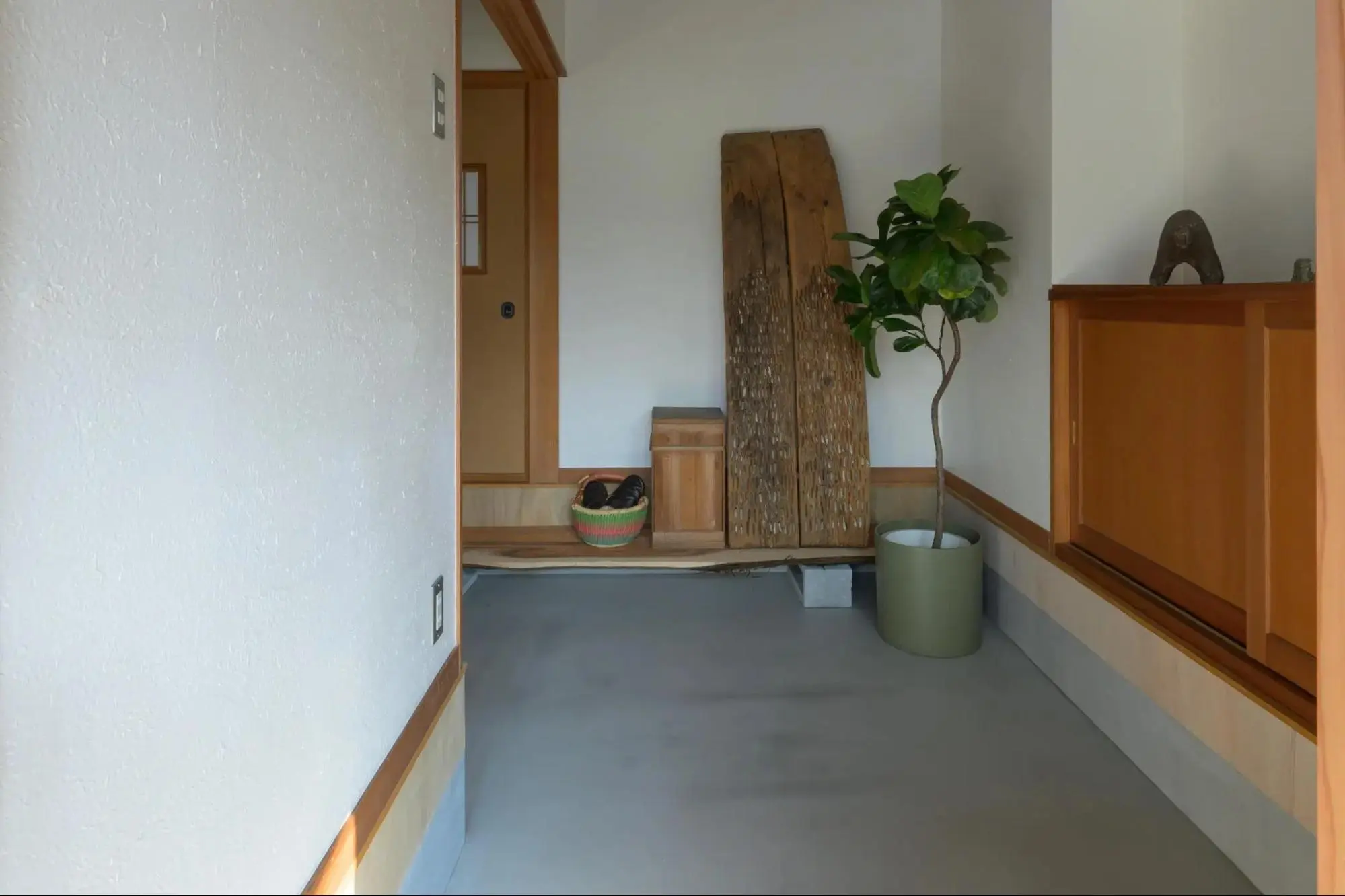 福井の戸建てフルリノベーション後。玄関から続く広いグレーの土間空間。右側には自然な木目と皮を活かした上がり框があり、古材の板や観葉植物、竹かごが並ぶ。昭和の趣を残した和モダンな集いの場。EUGUE RENOVATIONのリノベーション事例。