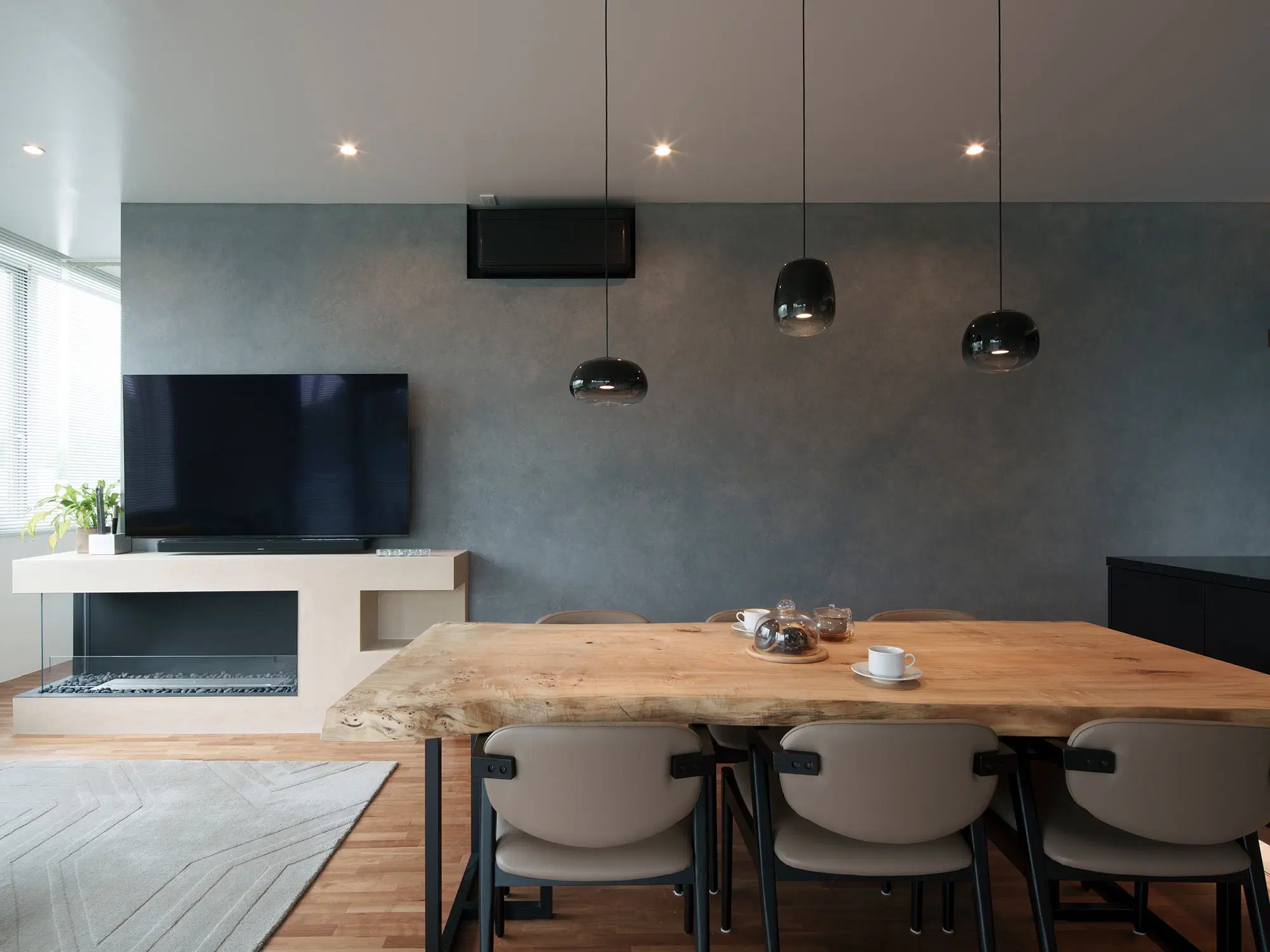 グレーの左官壁があるダイニングリビング。一枚板の木製テーブルの上には黒いペンダントライトが3つ吊り下げられ、左手にはテレビと暖炉が埋め込まれた棚が設置されている。
