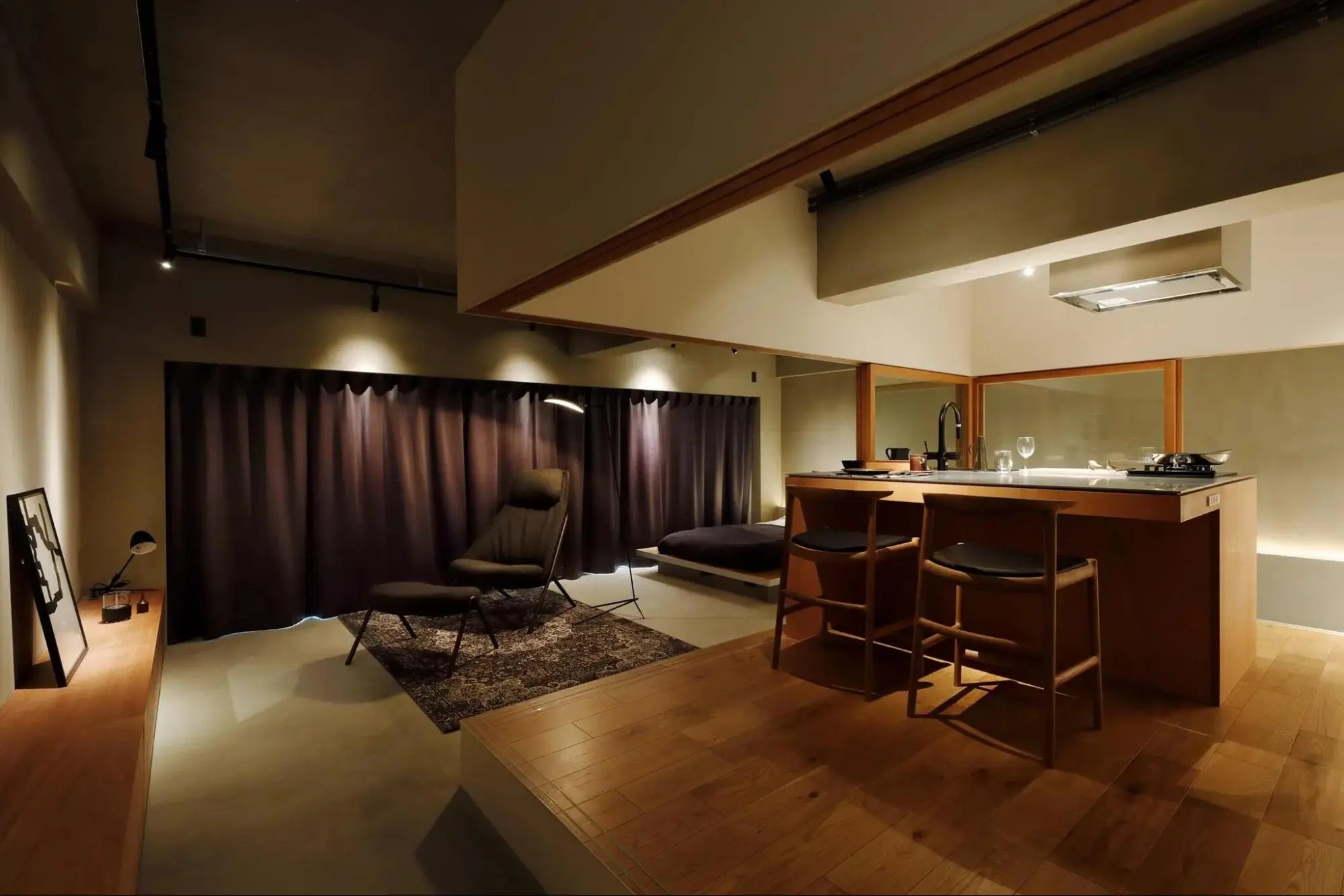 照明を落としたLDKの写真。手前に木製のアイランドキッチンカウンターとバースツールが置かれ、奥のリビングにはラウンジチェアが配置されている。