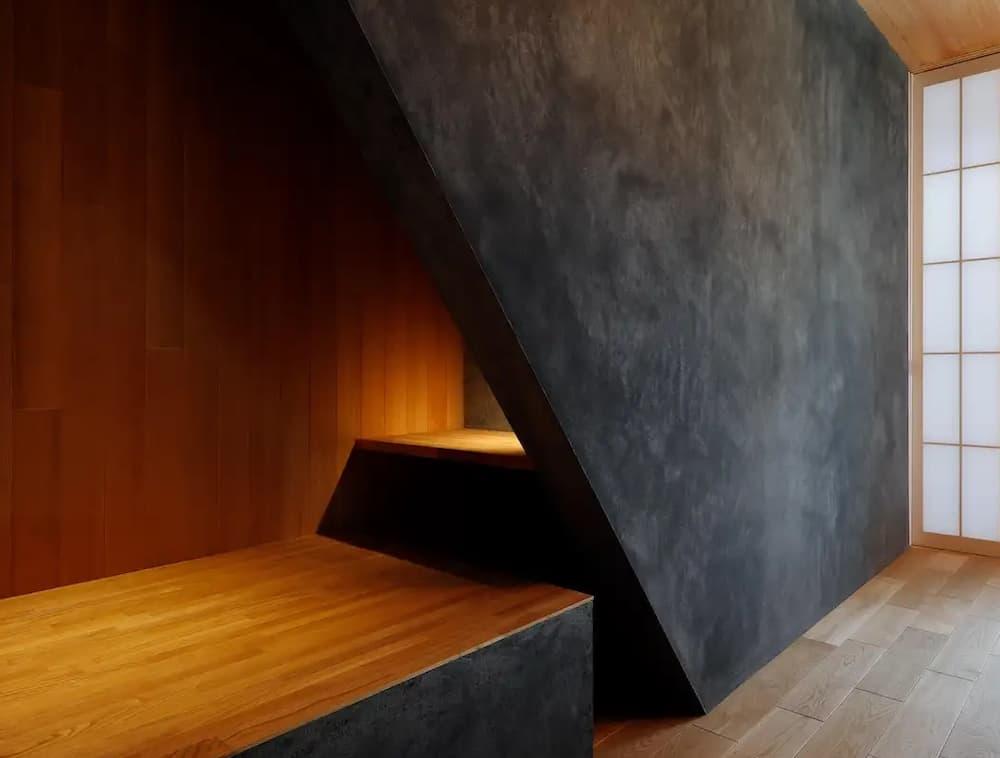 階段下の斜めのラインを活かし、入り口を鋭角に切り取った独創的なデザインのヌックスペース。外側の壁は重厚感のあるダークグレーの塗り壁（漆喰やコンクリート風）で仕上げられ、内部には温かみのある明るい色調の木目パネルが貼られている。内部に設置された間接照明が木の質感を柔らかく照らし、奥行きと「おこもり感」を演出。床面はフローリングと一続きの造作ベンチになっており、右奥には和の趣を感じさせる障子のような窓枠が見える。無駄な線を削ぎ落とした、静寂でモダンな雰囲気のインテリア空間。