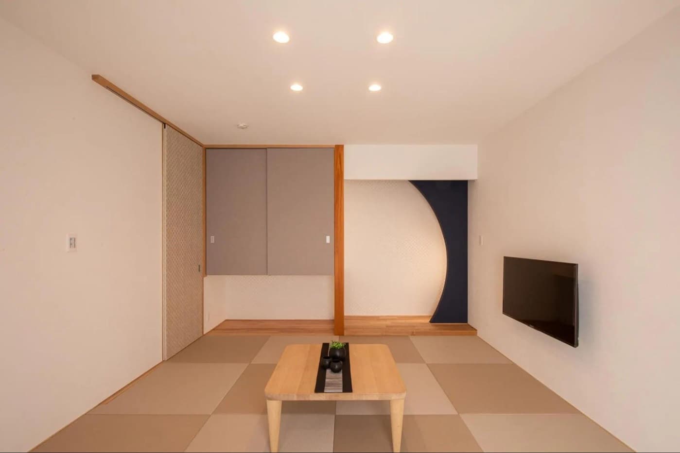 正方形の縁なし畳を敷き詰めたモダンな和室。正面にネイビーのアクセントクロスとアーチ開口の床の間、吊り押入れと間接照明を備える。