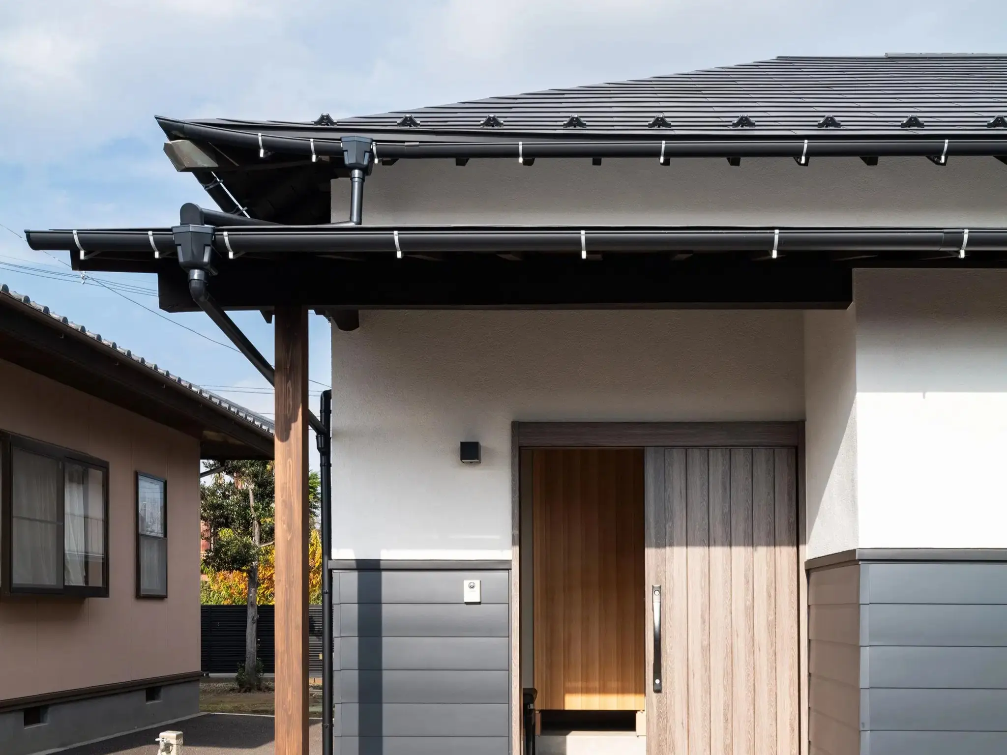 築80年の平屋を和モダンテイストにフルリノベーションした住まいの外観。白を基調とした上部の外壁と、ダークトーンのサイディングで仕上げた腰壁のコントラストが、建物の水平ラインを美しく引き立てている。玄関には温かみのある木製引き戸を採用。深い軒を支える立派な木柱が、古民家が刻んできた歴史と趣を色濃く残している。現代の意匠と建物の記憶が美しく調和した、株式会社AS IT ISのフルリノベーション事例。