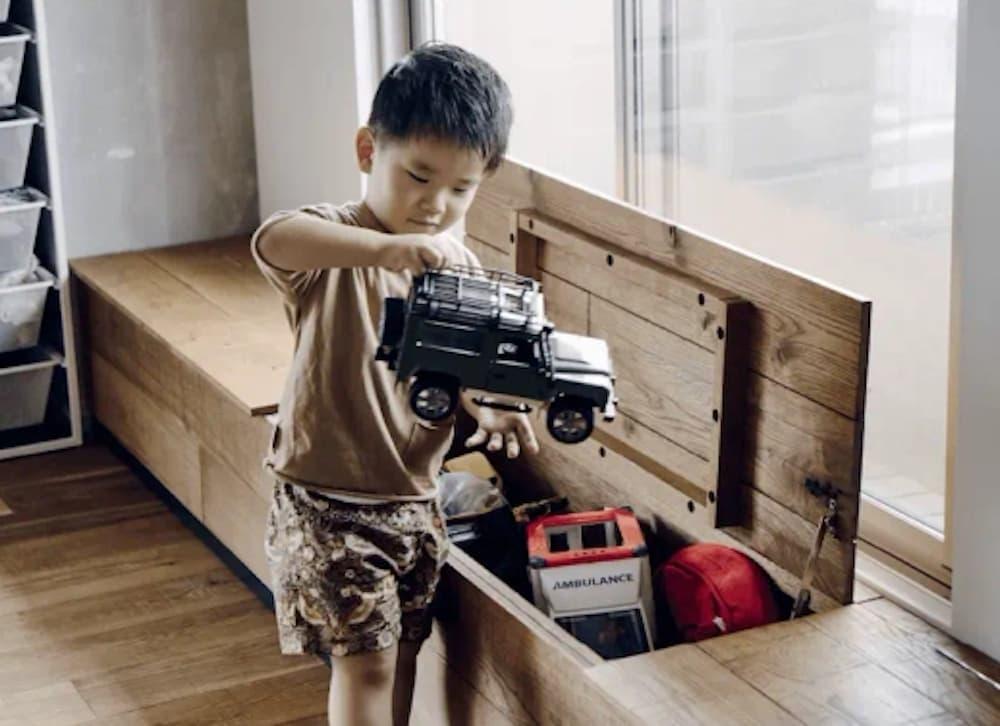 明るい光が差し込む大きな窓際に設けられた、温かみのある無垢材の造作ベンチヌック。座面が跳ね上げ式の蓋になっており、内部にはおもちゃをたっぷりしまえる収納スペースが確保されている。男の子が黒いオフロードカーの玩具を手に持ち、ベンチ下の収納に入っている救急車などの玩具と一緒に遊んだり、片付けたりしている。フローリングと調和した素材感と、生活動線に配慮した機能性を兼ね備えたリノベーションの実例写真。