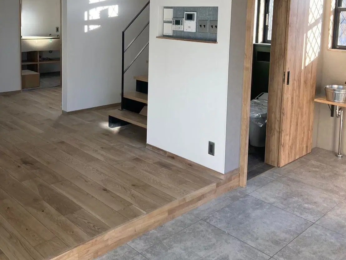 手前はグレーのタイル敷きの土間スペースで、木目調フローリングのリビングへ繋がる。リビング奥には黒いアイアン手摺のスケルトン階段を配置。壁面にはスイッチ用のニッチ、右手には木目引き戸のトイレがあるナチュラルな空間。an cube(アン キューブ)のリノベーション事例。