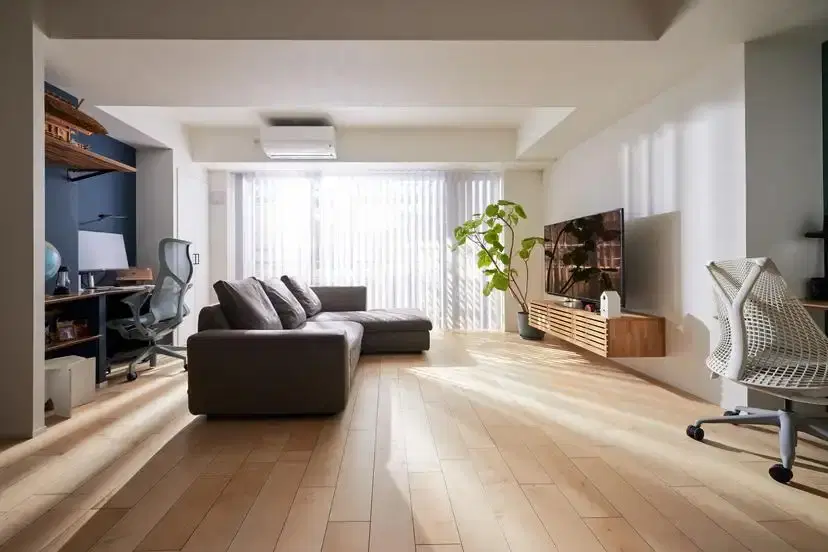 縦型ブラインドから自然光が入る、木目フローリングのLDK。中央にグレーのL字ソファ、壁面には木製の浮き型テレビボードを配置。左奥にはブルーのクロスが特徴的なワークスペースと棚が設けられている。empatiの施工事例。