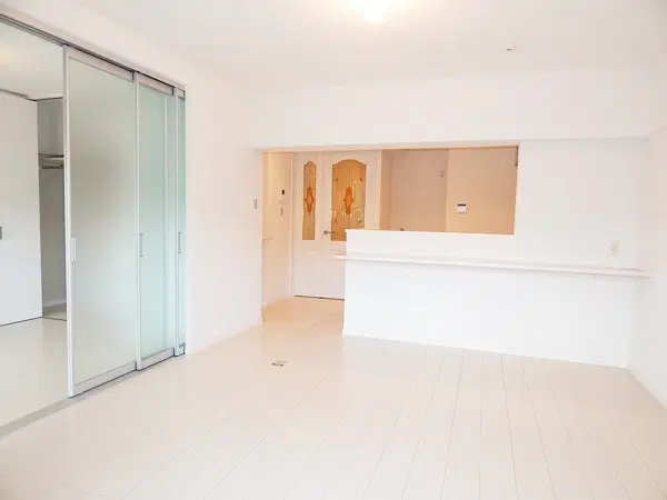 リノベーション後の、白を基調とした明るいLDK。床は白い木目調で、左手には半透明ガラスの引き戸、奥にはキッチンの腰壁と装飾ガラスのドアがある。