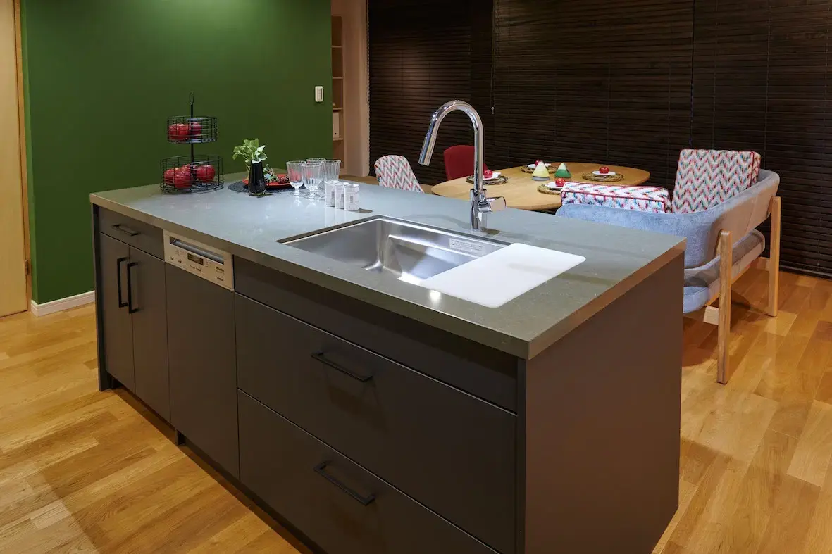 中央にダークグレーのキャビネットとグレー天板のアイランドキッチン。ステンレスシンクと水栓、フルーツ。背後はグリーンのアクセントウォールと木製ルーバー。木目フローリングのモダンなダイニングキッチン。株式会社ディセノ施工。