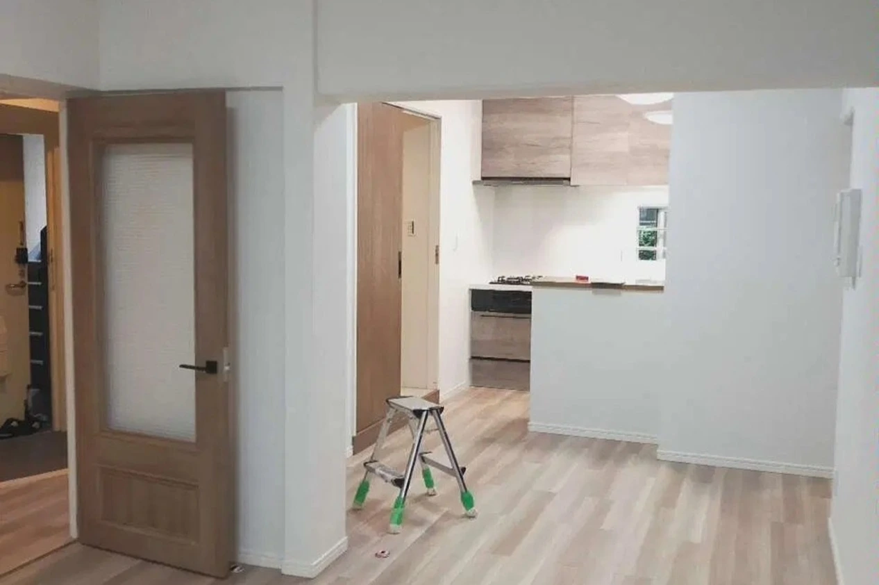 白いクロス貼りの壁と大きな梁で構成された、fujitacaリノベーションのリノベ前内観。奥にはセミクローズド型のキッチンがあるが、垂れ壁や袖壁により閉鎖的な印象。手前には木製ドアと脚立があり、構造壁の圧迫感を解消する前の既存状態。