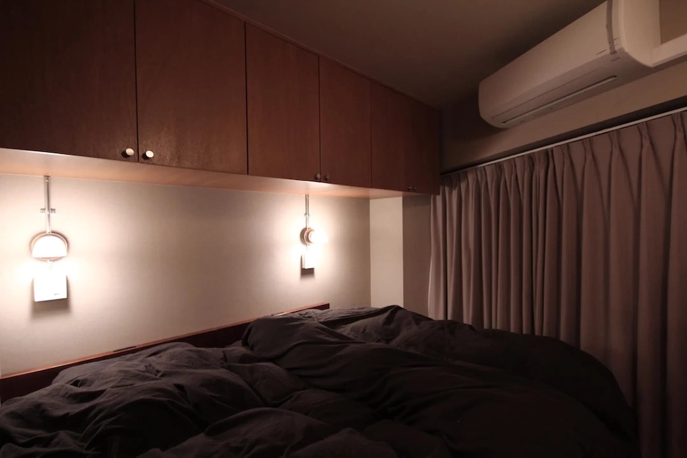 ダークブラウンの木目調吊り戸棚を壁一面に設置し、収納力とデザイン性を両立させた大人シックなインダストリアル寝室。ダークグレーのベッドリネンとグレージュのカーテンが落ち着いたトーンで統一され、枕元のレトロなブラケットライトが柔らかな光を灯す、ホテルライクで安眠を誘うシンプルモダンなベッドルームインテリア。