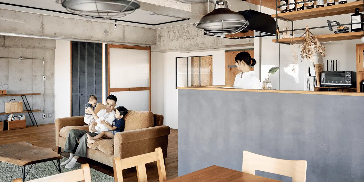 マンションフルリノベーション後のLDK。天井と左側の壁はコンクリート現し仕上げで、露出配管を施している。右側にはグレーのモルタル調腰壁の対面キッチンがあり、上部にアイアンと木材を組み合わせた吊り棚を設置。天井のダクトレールからは金属製のペンダントライトを吊り下げている。中央の木目調フローリングにはグリーンのラグを敷き、ブラウンのソファとアイアン脚の一枚板ローテーブルを配置。奥の壁には黒いルーバー扉、木枠の引き戸、黒枠の室内窓を設けている。株式会社スクエアのリノベーション事例。