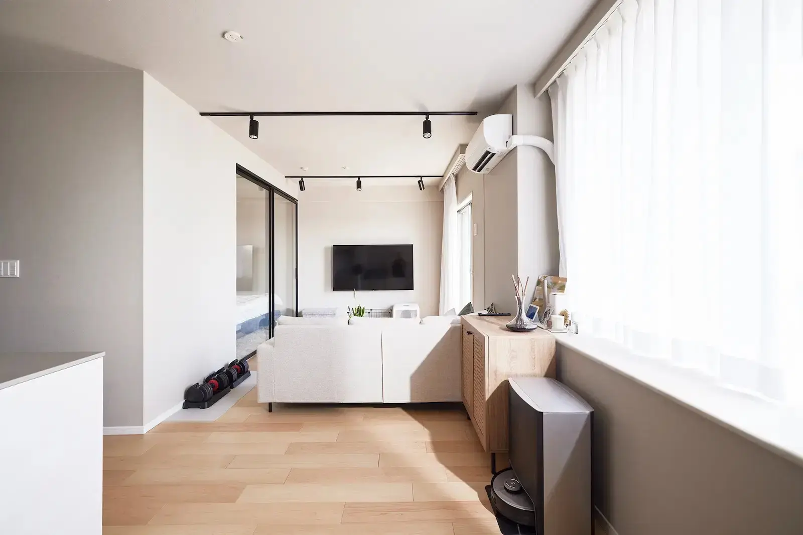 明るいヘリンボーン床のLDK。奥にグレーの対面キッチンとダイニングセットがあり、手前のリビングにはソファとテレビがある。ソファ背面にガラスの間仕切りが見える。