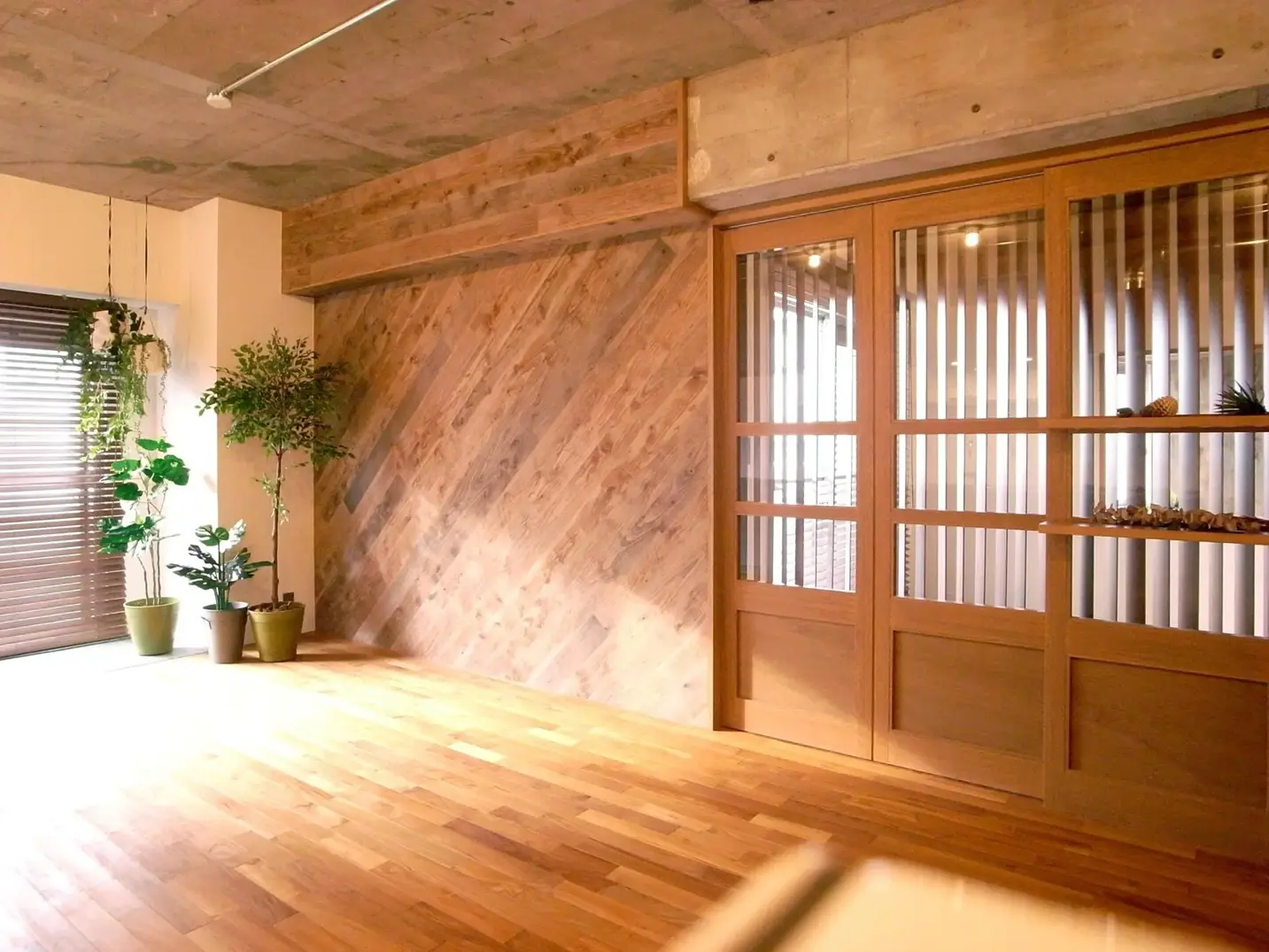 コンクリート現しの天井と、斜め張りの天然木パネルの壁が特徴的な空間。床材にはチークを使用し、右側には格子デザインの造作ガラス引き戸を設置。窓際には観葉植物が置かれた、ラスティックなマンションリノベーション。田中建設の施工事例。