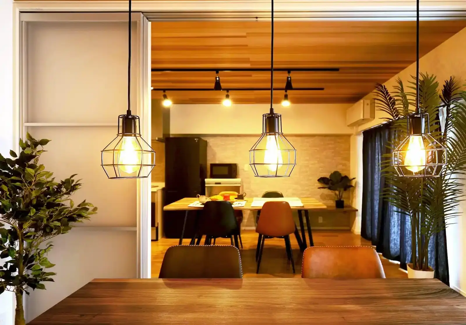 印象的なワイヤーフレームのペンダントライト越しに広がる、カフェのようなダイニングキッチン。木目調の天井にブラックのダクトレール照明を合わせ、インダストリアルで温もりのある空間を演出したリ・ワークショップの施工事例。