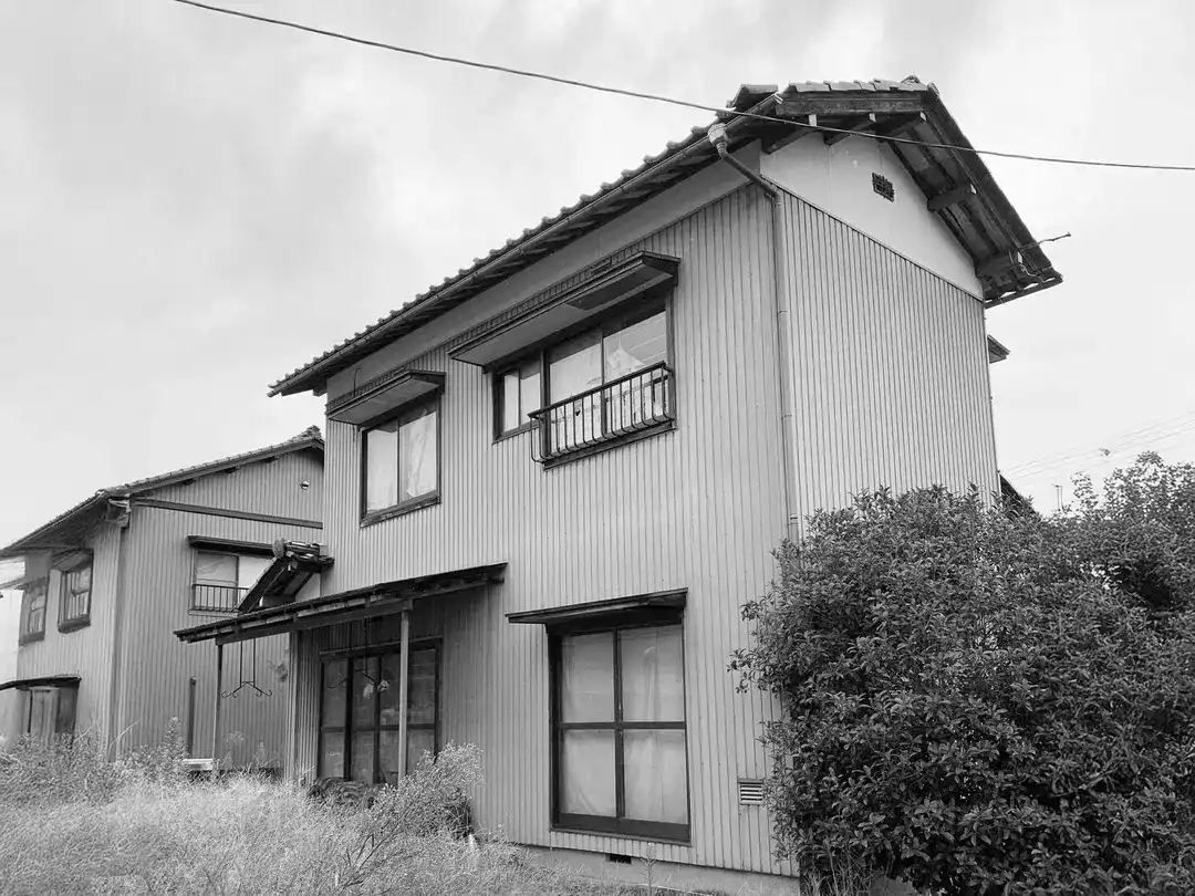 リノベーション前の古い木造住宅の外観。白黒写真で、壁は縦張り、庭には草木が茂っている。