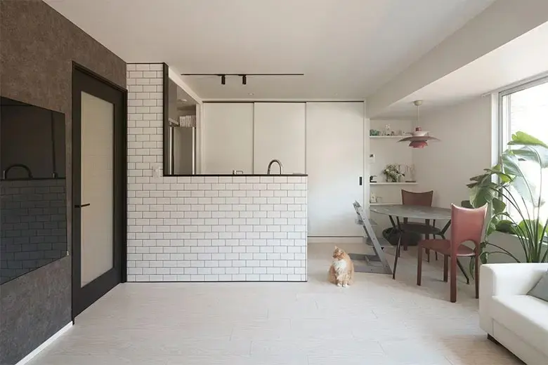 白いサブウェイタイルを貼ったキッチンカウンターがあるLDK。床は白系のフローリングで、茶色の猫が座っている。左手には黒いフレームのドアが見える。