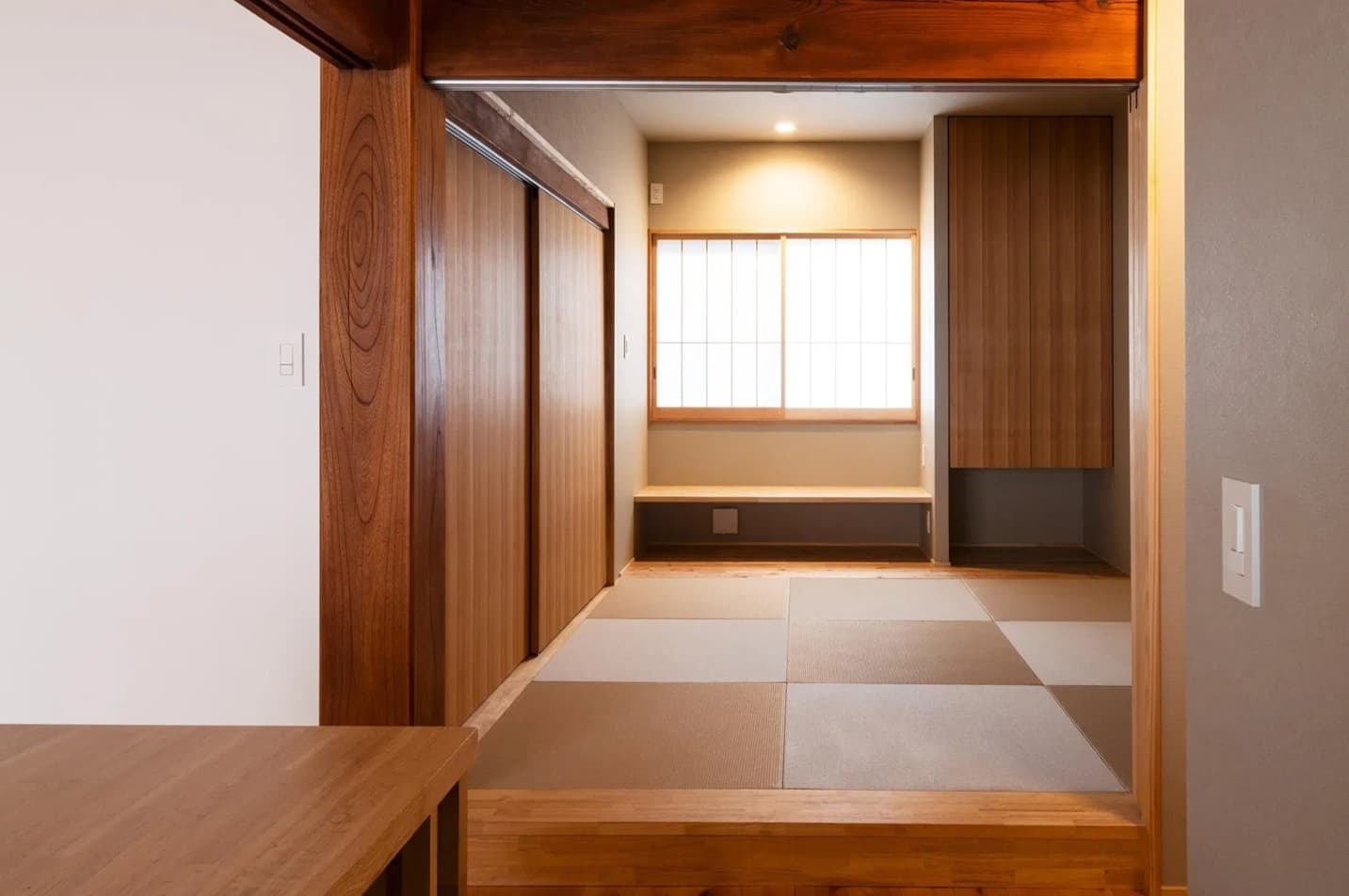 太い既存の柱と鴨居が残る、一段高くなった和室スペース。市松模様に敷かれた縁なし畳、障子窓、木製の造作収納が配置された、古民家の趣を感じる落ち着いた内観。