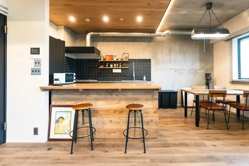 ダイニングキッチンの写真。古材のカウンターにバースツールが置かれ、壁は黒いサブウェイタイルとコンクリート。奥にダイニングセットがある。