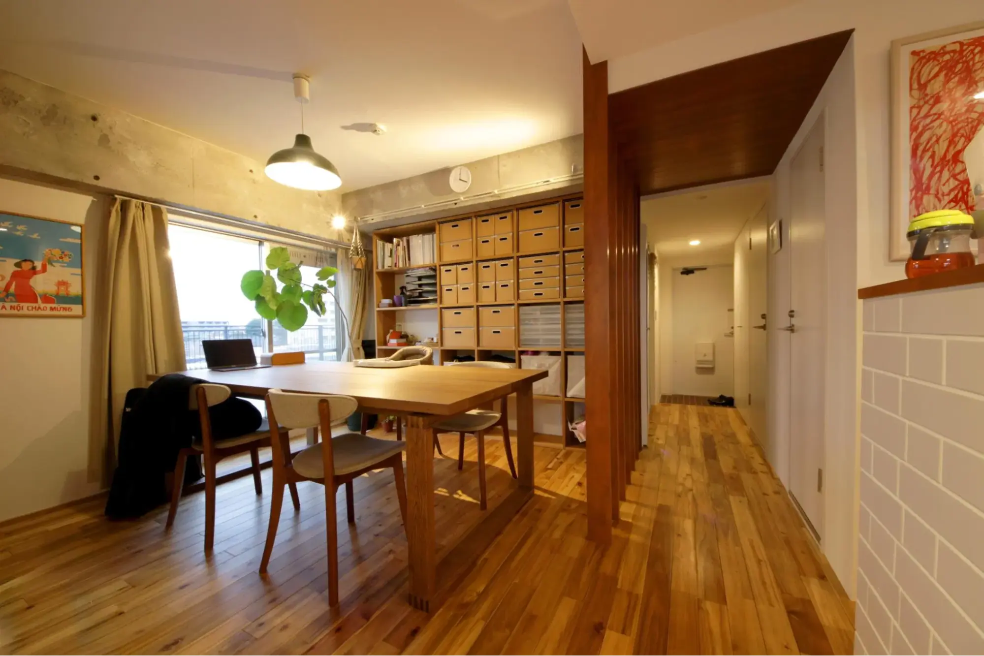 温かみのある無垢材フローリングのダイニング空間。中央に大きな木製テーブル、奥には天井まで届く大容量の木製オープン棚を配置。窓上のコンクリート現しの梁や、廊下をゆるやかに仕切る木の縦ルーバーが、カフェのような心地よい空気感を演出している。株式会社田中建設のリノベーション事例。