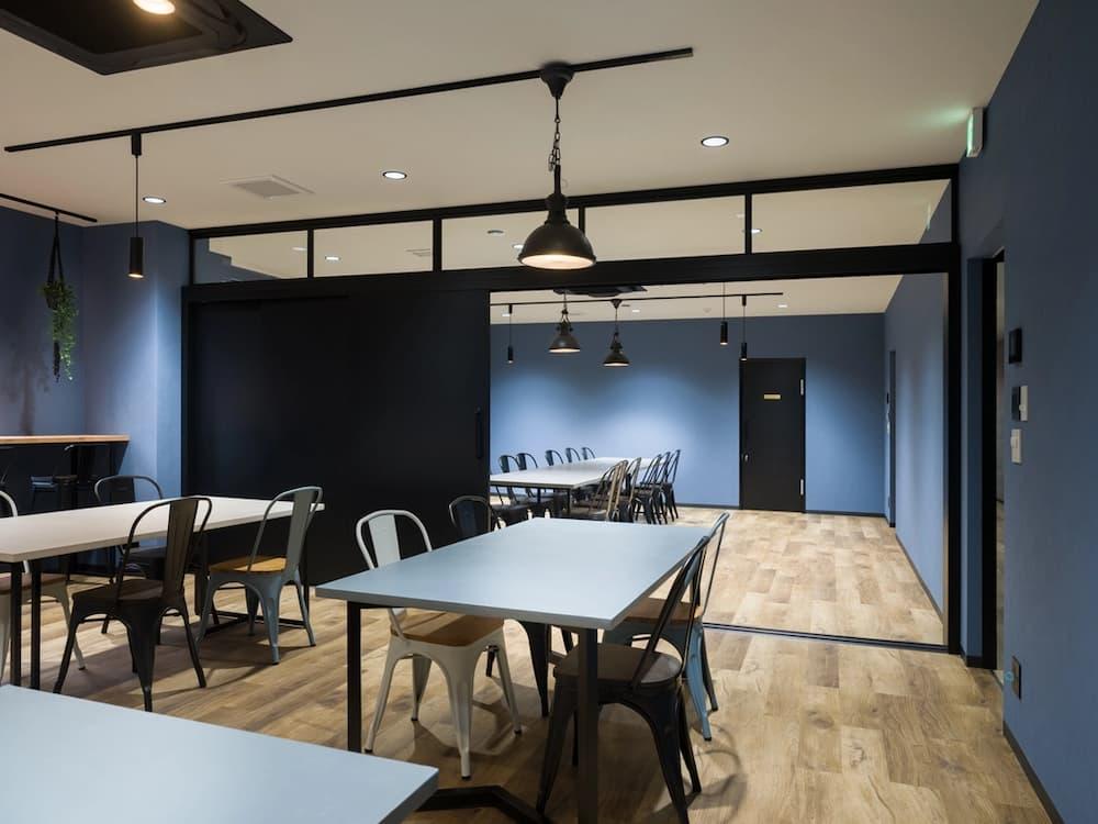 深ブルーの壁紙と木目調の床で構成されたカフェテリア兼会議スペース。黒い鉄枠のガラス間仕切り戸で空間を区切り、工業用照明やスチール製の椅子を配置している。金属加工工場のオフィスらしく、鉄と木を組み合わせたインダストリアルな内装デザイン。
