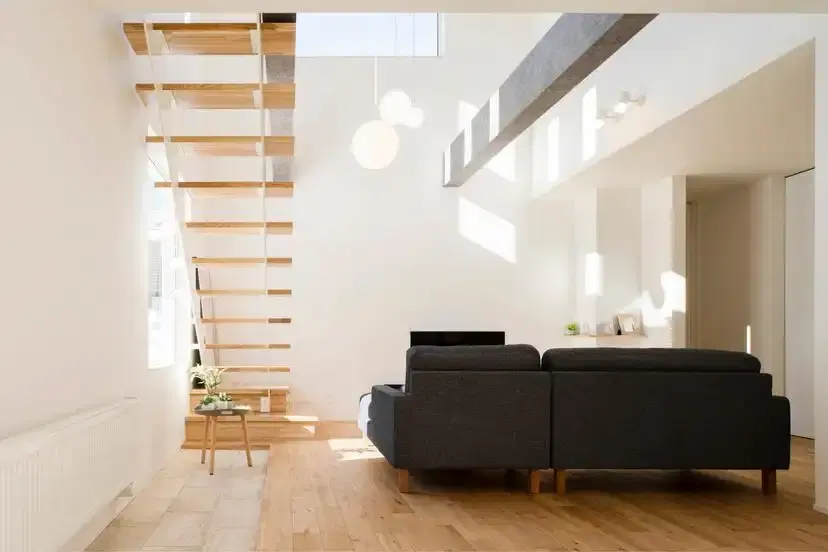 吹き抜けの高い天井から自然光が差し込む、白を基調としたLDK。無垢材の踏板を使用したスケルトン階段が壁際に配置され、中央にはダークグレーのローソファが置かれている。余白を活かしたミニマルで開放感のある戸建てリノベーションの内観