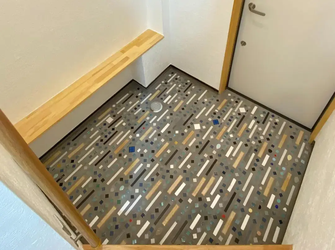 グレーの玄関土間床に、長方形や正方形の多色タイルがランダムに多数埋め込まれている。左手には木製の造作ベンチ、右奥には白い扉を配置した空間。スケールメイクスのリノベーション事例。