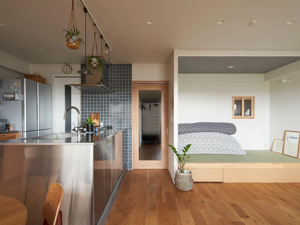 ステンレス製のスタイリッシュなアイランドキッチンの隣に広がる、畳（タタミ）を敷いた小上がりのヌックスペース。ベース部分は明るい木目調で、下部には便利な引き出し収納が備わっている。ヌックの天井は落ち着いたダークグレーで一段低く設計されており、白い壁とのコントラストが心地よい「おこもり感」を演出。内部には大きなグレーのクッションや立てかけられたアート、小さな木枠の内窓があり、リラックスした雰囲気が漂う。リビングのフローリングと一続きになりながらも、段差と天井の色使いによって独立した休息空間として定義された、機能美あふれるインテリア。
