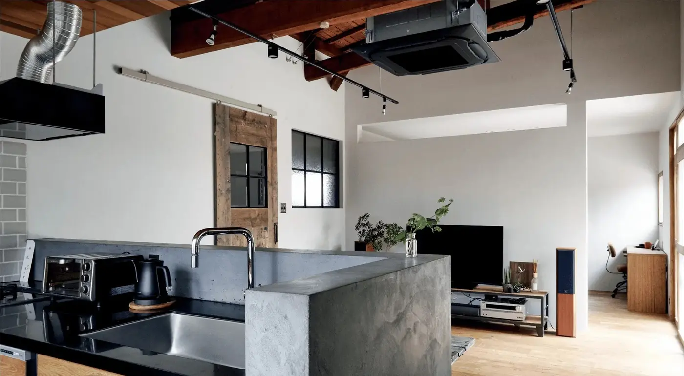 モルタル仕上げのキッチン腰壁と、既存の梁や小屋組を現しにした高い天井が特徴的なLDK。ヴィンテージ風の木製ドアや黒枠の室内窓、露出配管がブルックリンスタイルを演出する、築60年の古民家リノベーション内観。株式会社スクエアの施工事例。