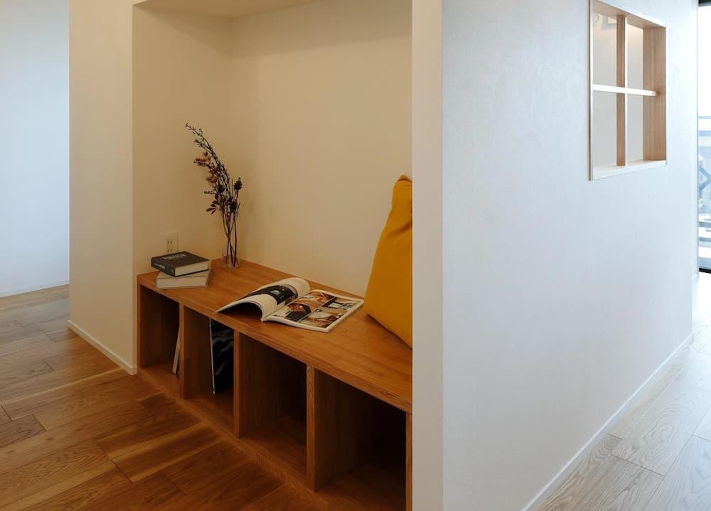 白壁の通路の一角を凹ませて作った、秘密基地のような「こもり感」のある造作ベンチヌック。明るい色味の無垢フローリングと馴染む木製のベンチ下には、本や雑誌をしまえるオープンな収納スペースを確保している。ベンチの上には黄色いクッションや読みかけの本、ドライフラワーが飾られ、壁面にはスマホの充電に便利なコンセントも設置。右側の壁には小さな室内窓が設けられ、閉鎖的になりすぎず家族の気配も感じられる、廊下やデッドスペースを有効活用したリノベーション事例。