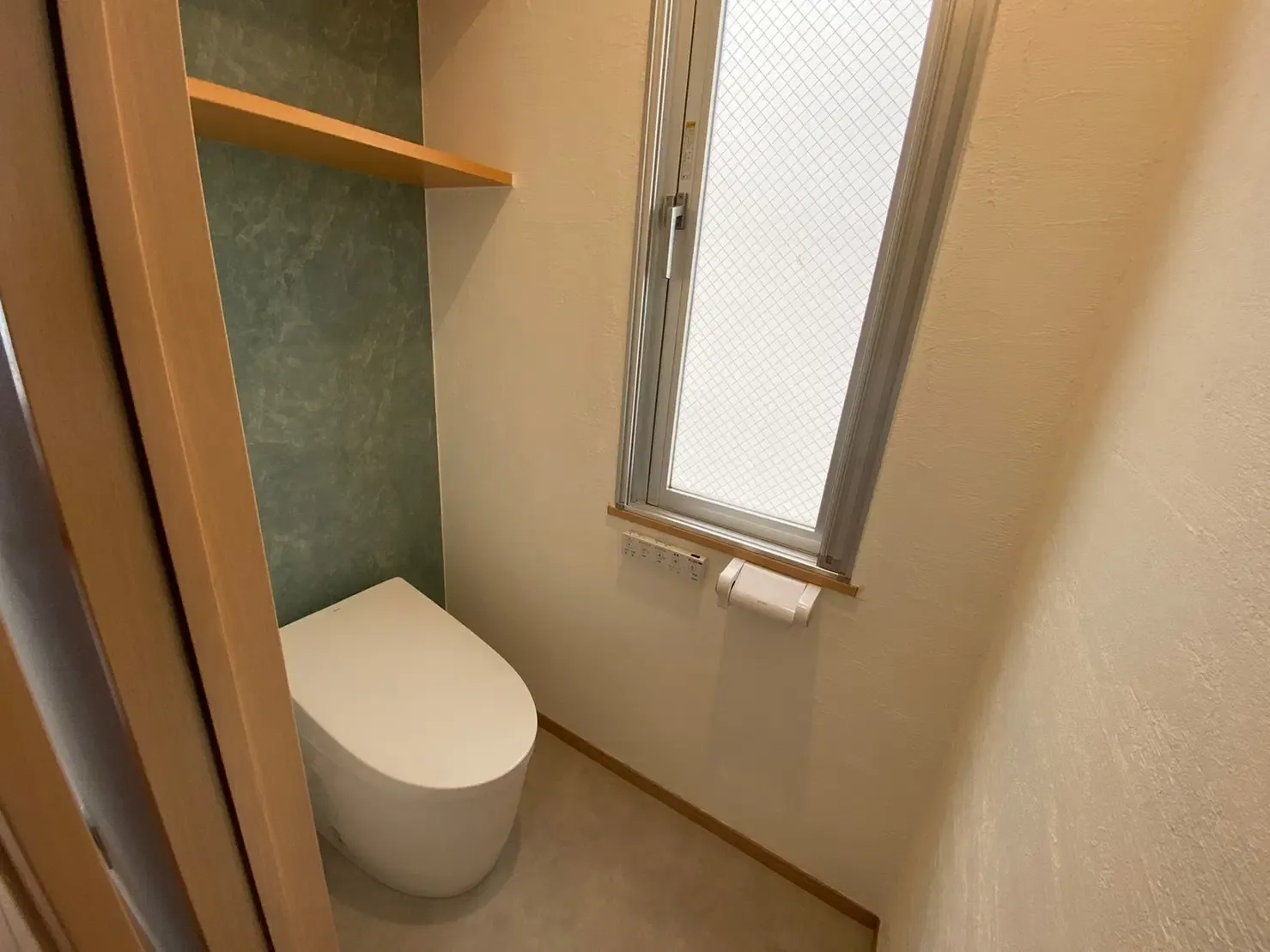 戸建てトイレのリノベーション後。手前に白いタンクレストイレを配置。壁面は無添加漆喰仕上げで、トイレ背面のみグリーンのアクセント壁となっている。背面上部に木製の固定棚、右手の壁に縦長の窓とリモコン、ペーパーホルダーを設置している。有限会社スケールメイクスのリノベーション事例。